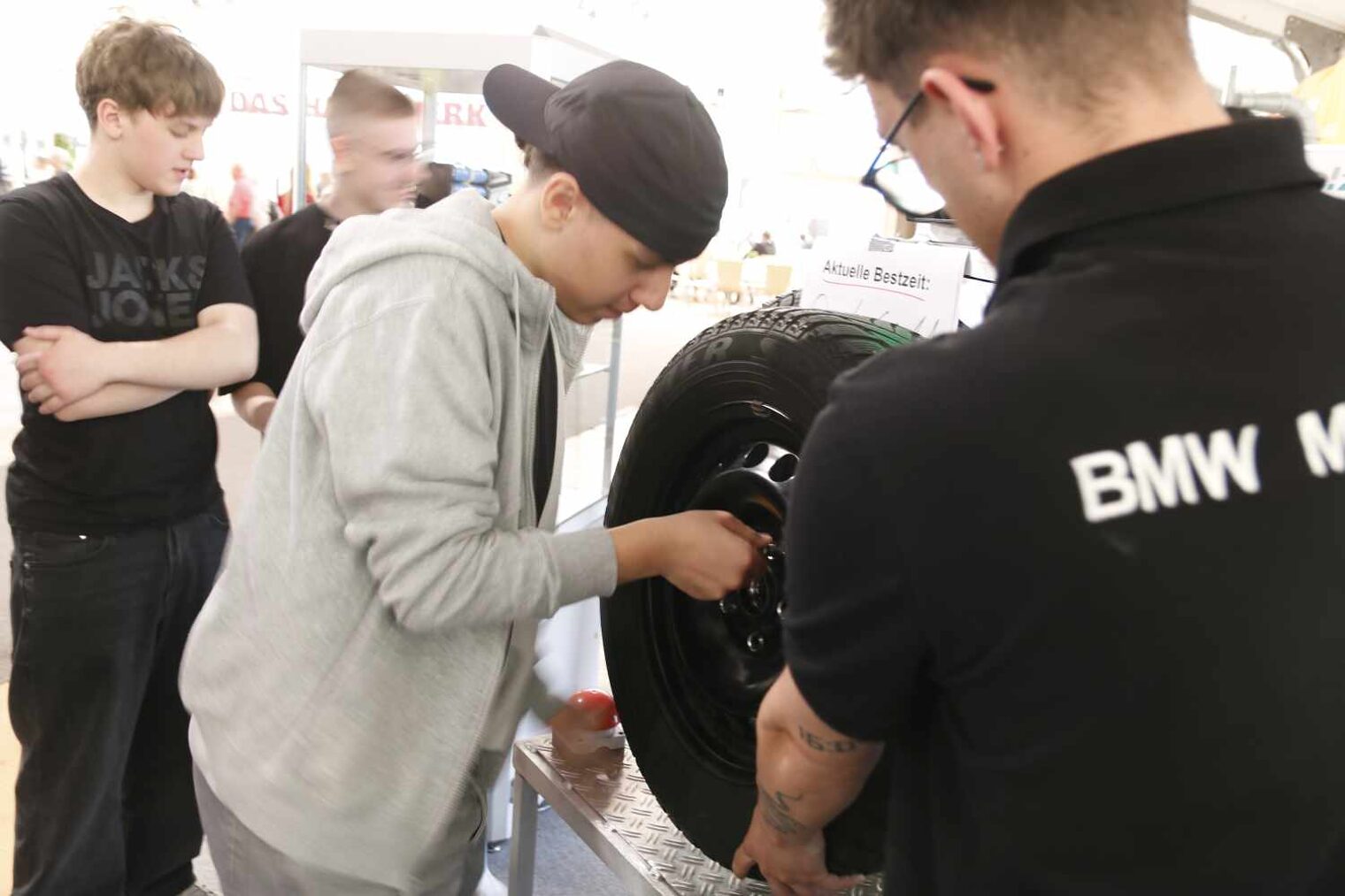 Junger Mann mit grauem Hoody und schwarzem Cap schraubt einen Reifen an ein Gestell, im Vordergrund der Rücken eines Mannes, der den Reifen hält, hinten stehen 2 Jungs