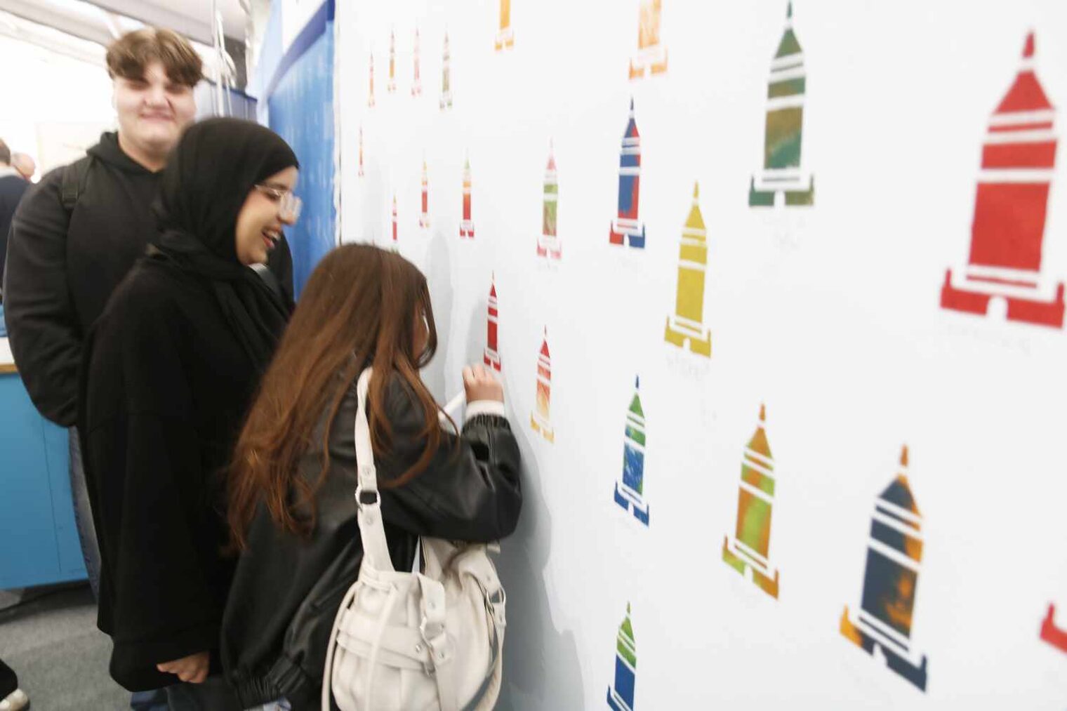 Drei Personen in der linken Bildhälfte an einer weißen Wand mit bunten Turmmotiven, die die rechte Hälfte ausfüllt. Der Junge schaut in die Kamera, die beiden Mädels malen etwas.