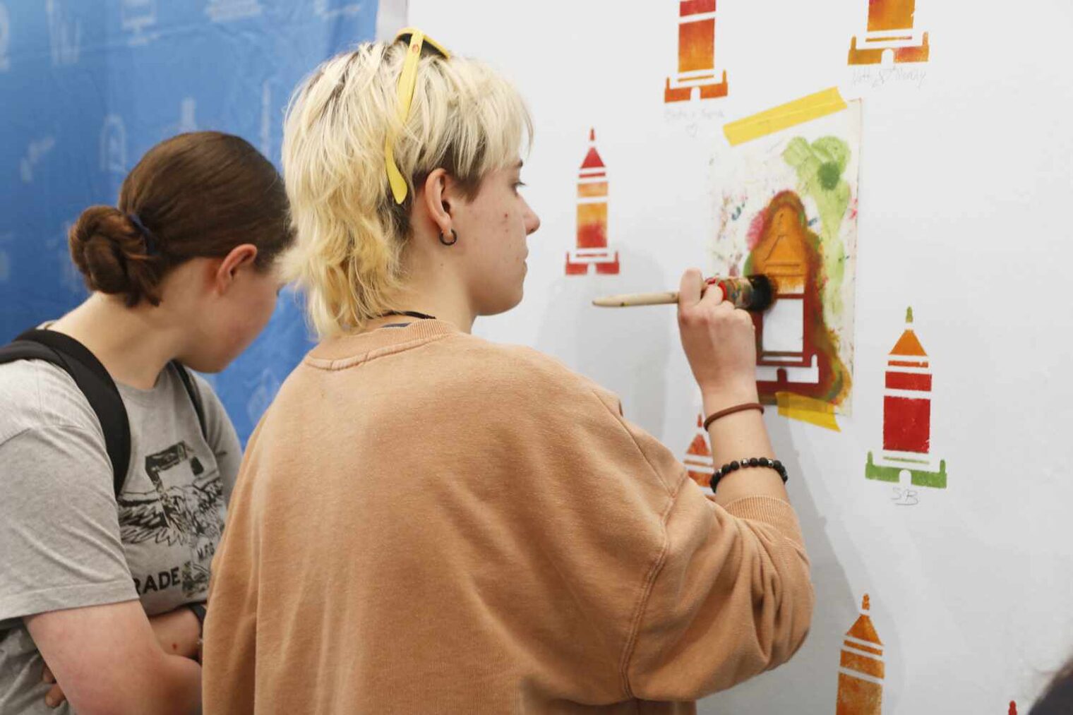 Jugendliche mit kurzen blonden Haaren und weitem beigen Pulli tupft über eine Schablone in Turmform Farbe an die weiße Wand, dahinter schau ein anderes Mädchen zu Boden
