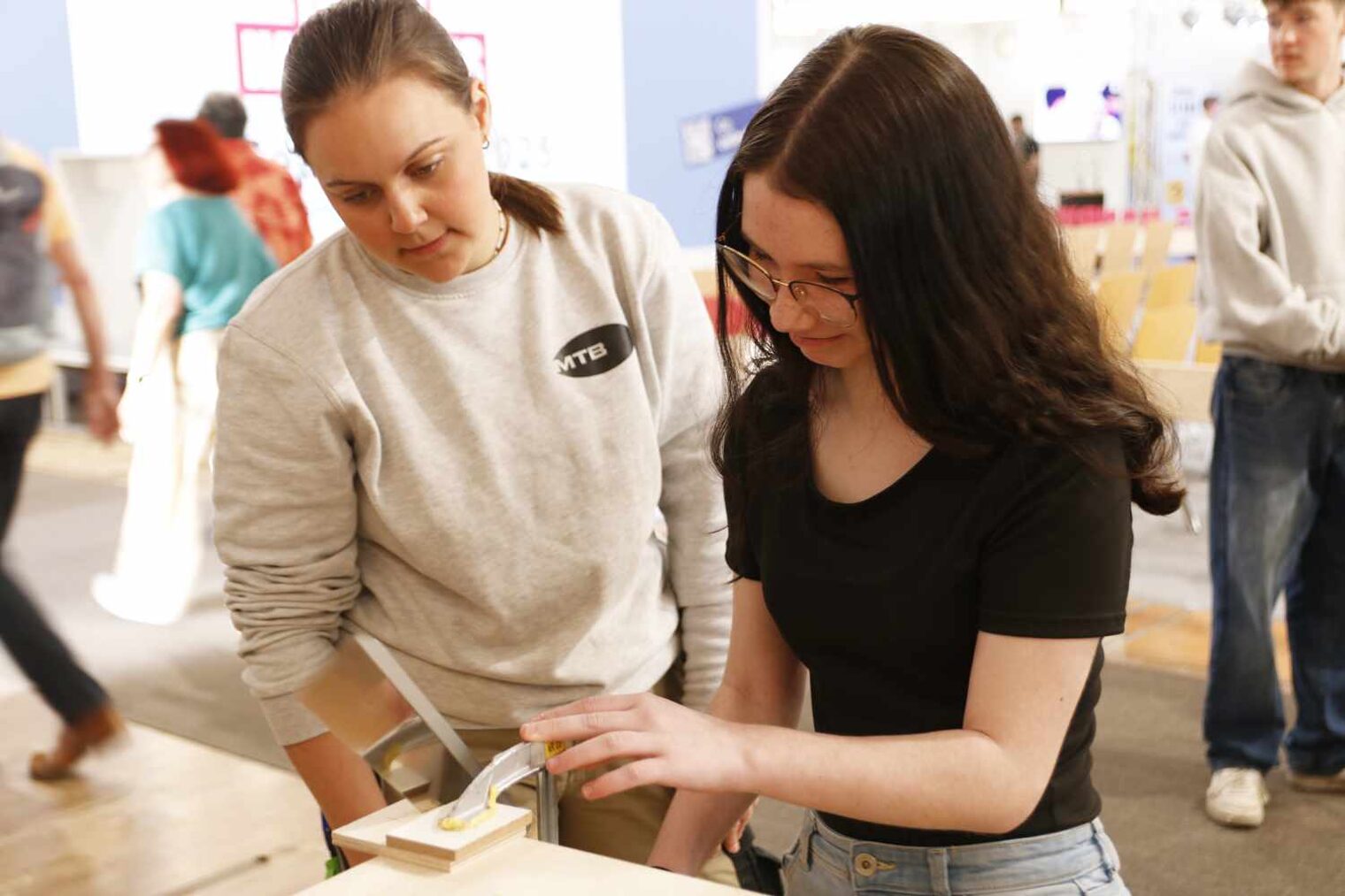 Zwei junge Frauen an der Ecke eines Arbeitstisches, an den ein Holzstück eingespannt ist, die rechte Frau arbeitet daran, die linke Frau blickt nach vorne gebeugt zu.