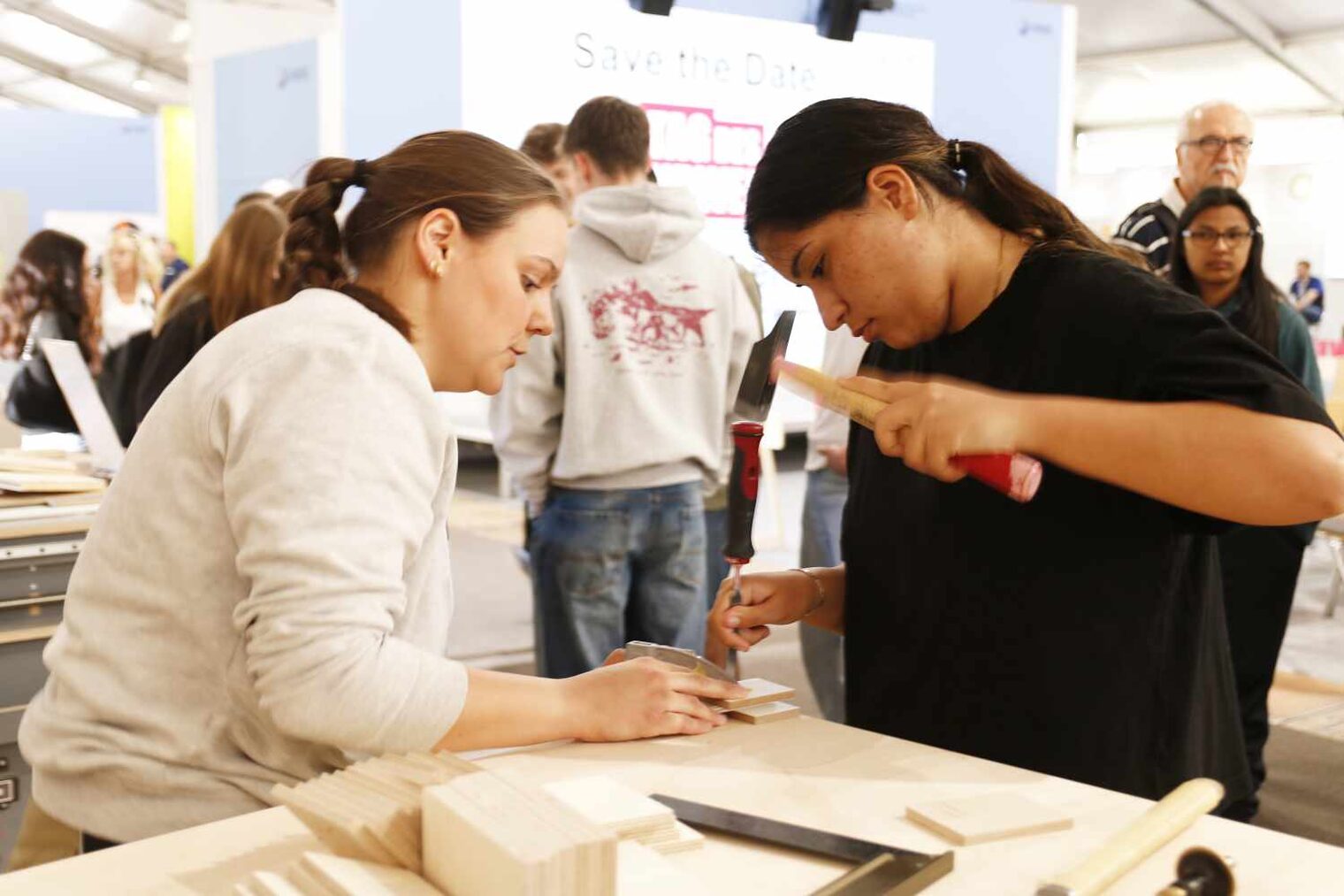An der Ecke eines Arbeitstischs, steht links eine Schreinerin, die ein Holzstück hält, rechts neben ihr eine junge Frau, die mit dem Hammer ausholt.