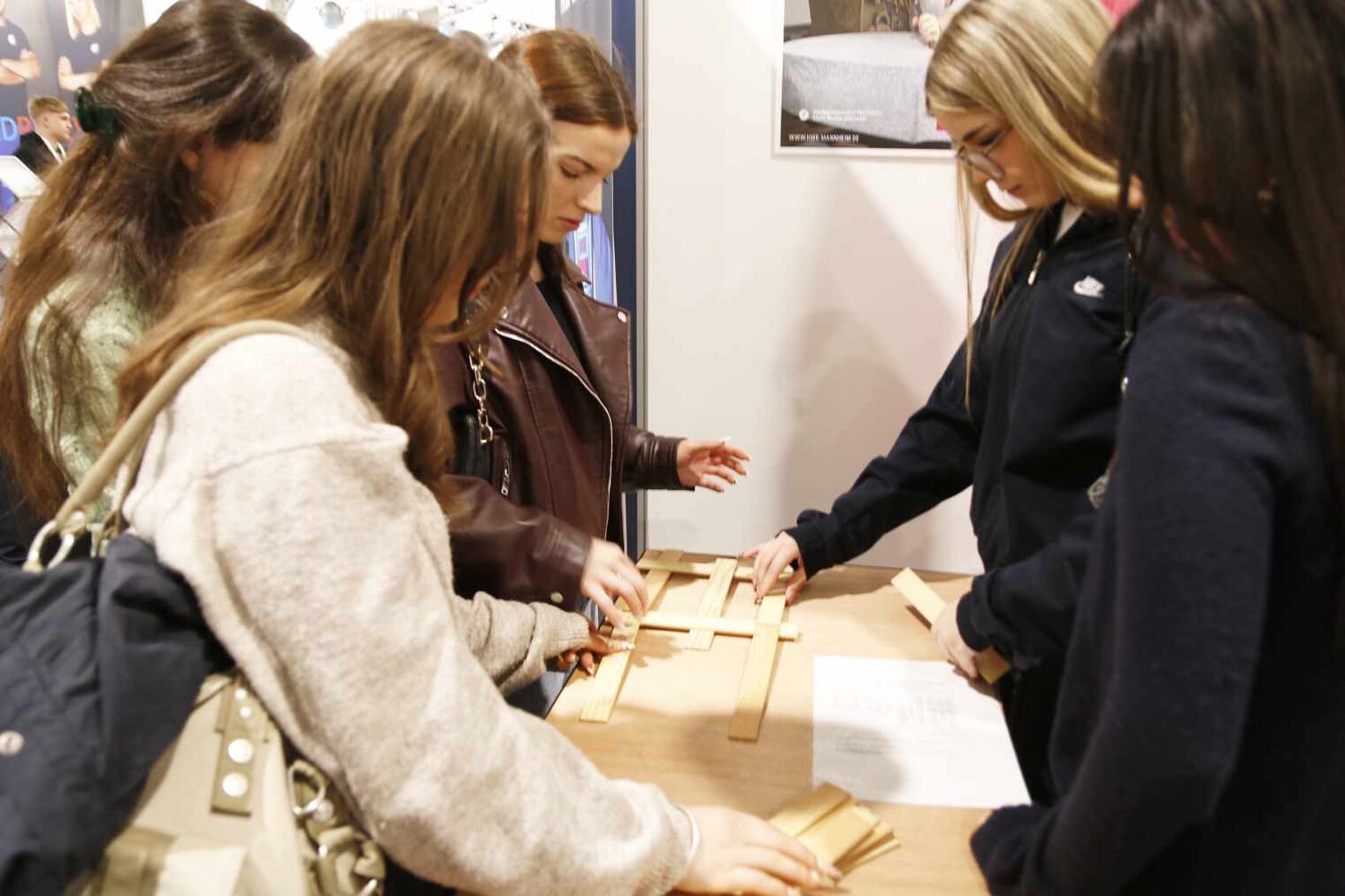Links drei und rechts gegenüber zwei junge Frauen an einem schmalen Holztisch, konzentriert nach unten schauend, legen kleine Holzstücke nach Plan übereinander