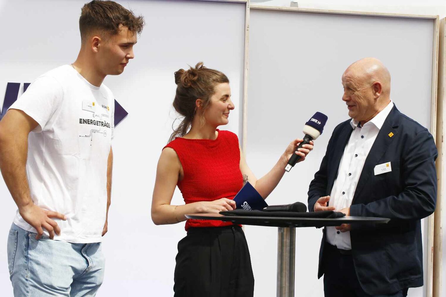 Drei Personen auf einer Bühne an einem Stehtisch, in der Mitte junge Frau im roten Top, die den Mann im Anzug rechts neben ihr interviewt, junger Mann links mit Händen in den Hüften