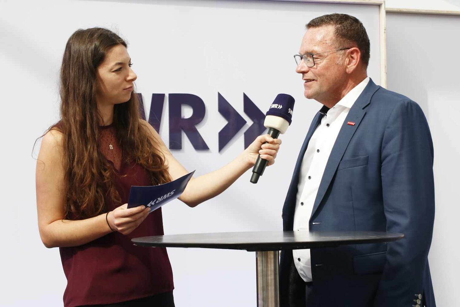 Frau mit langen braunen Haaren und Moderationskarte in der Hand interviewt mit Mikro in der anderen Hand den Mann rechts im Anzug, im Vordergrund ein Stehtisch