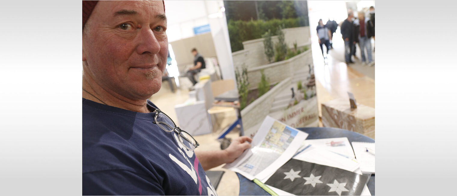 Mann am linken Bildrand blickt über die Schulter in die Kamera, vor ihm auf dem Stehtisch sind Unterlagen zu sehen, darunter ein schwarzes Papier mit silbernen Sternen