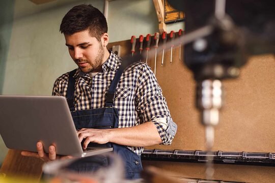Mann in kariertem Hemd hält Laptop in der Hand und im Vordergrund unscharf eine Bohrmaschine. Mann schaut auf Bildschirm. 