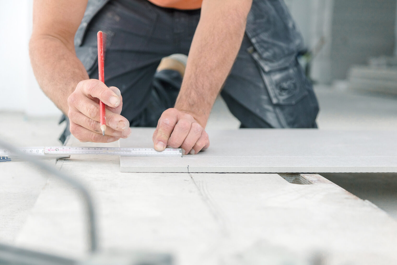 Ausschnitt eines Fliesenlegers bei der Arbeit, der auf dem Boden kniet und mit einem Bleistift und Metermaß in den Händen etwas anzeichnet
