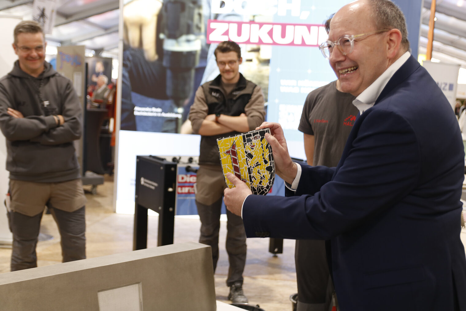 Mann mit dem Rücken zur Kamera zeigt ein aus bunten Fliesen gearbeitetes Wappen, im Hintergrund stehen zwei weitere Männer mit verschränkten Armen.
