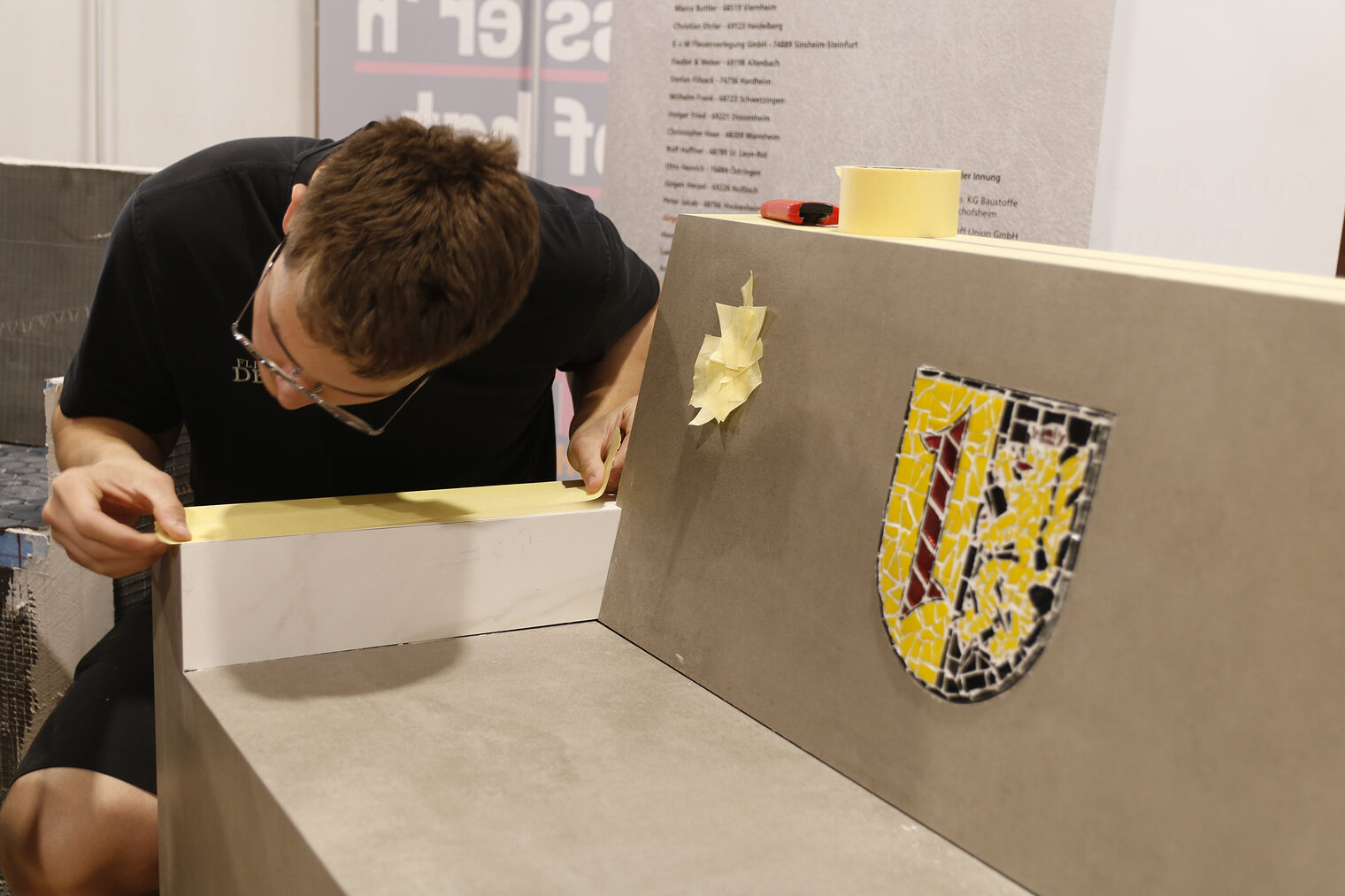 Junger Mann mit Brille beugt sich über die Lehne einer gefliesten Bank und spannt ein Kreppband, um es darauf zu kleben. In der Rückenlehne ein Wappen.