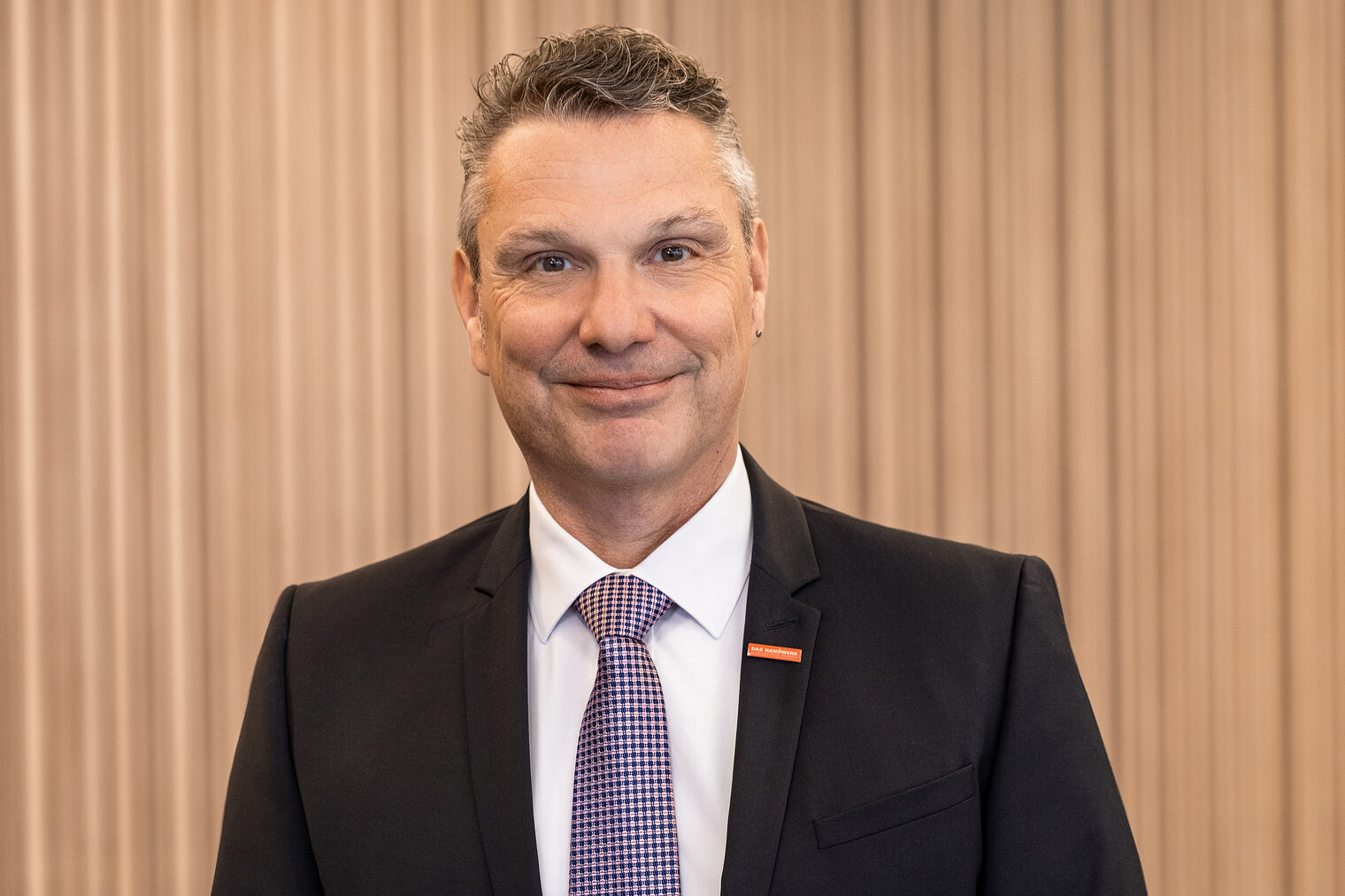 Mann mit grau melierten Haaren lächelt in die Kamera. Er trägt ein dunkles Jackett, weißes Hemd und eine fein karierte Krawatte.