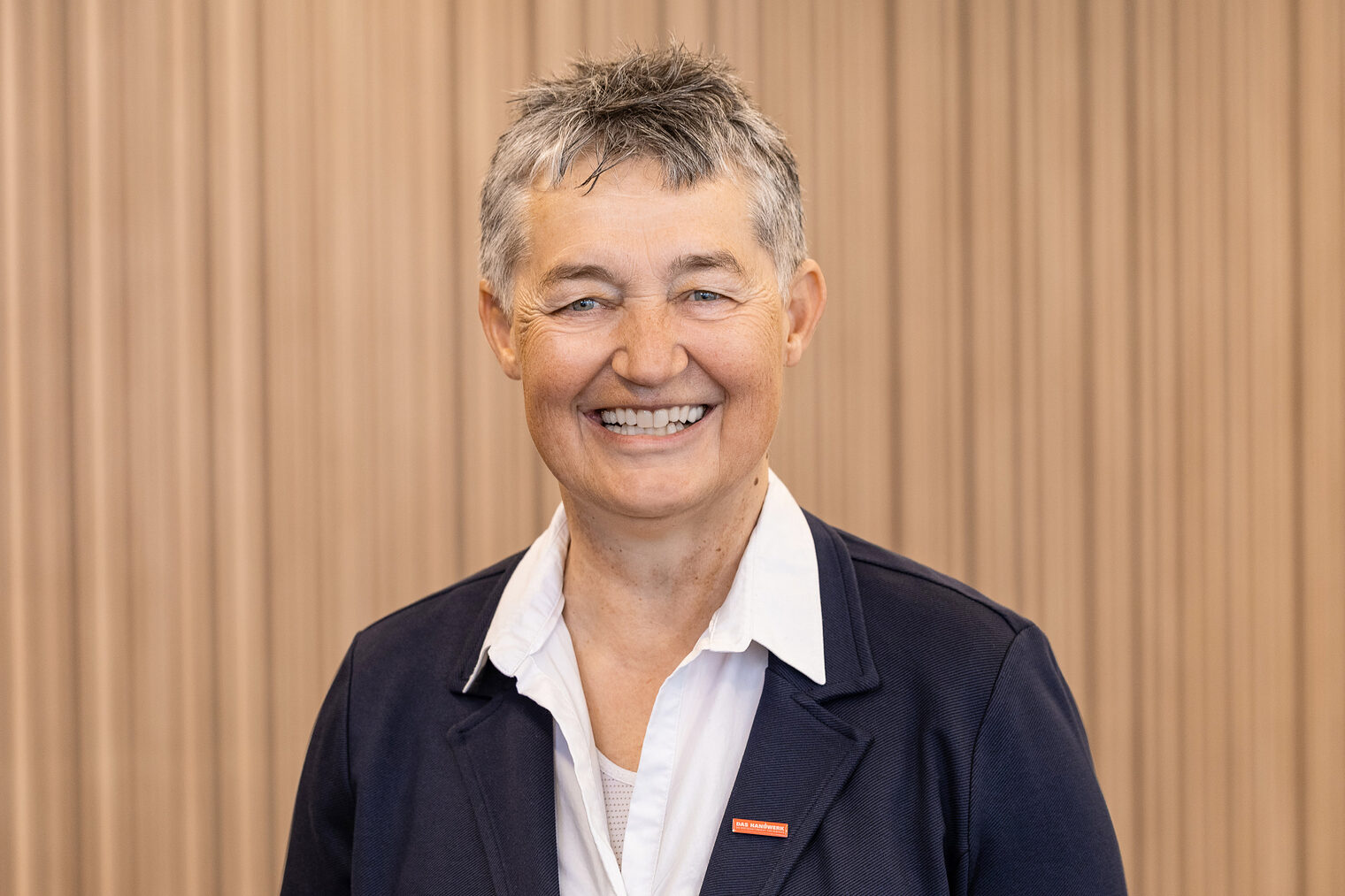 Frau mit kurzen, grau melierten Haaren lacht fröhlich in die Kamera, trägt dunkles Jackett mit rotem Anstecker am Revers und eine weiße Bluse mit leger geöffneten oberen Knöpfen.