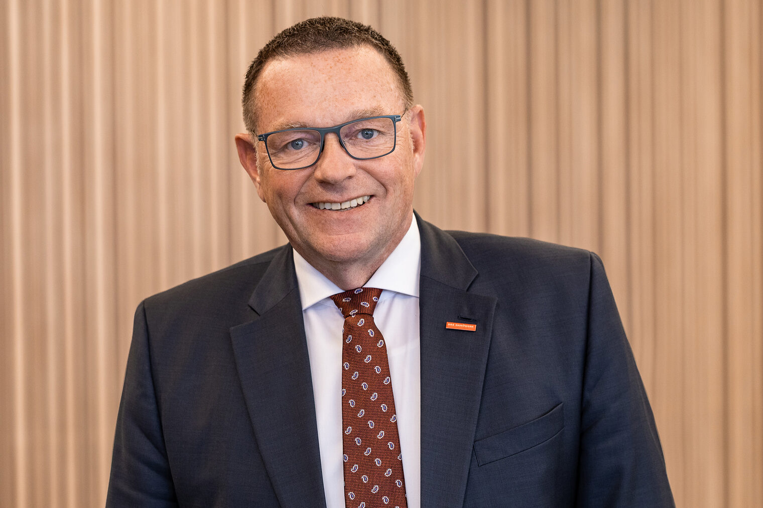 Mann mit grau gerahmter Brille und dunklen kurzen Haaren lacht in die Kamera, trägt dunklen Anzug, weißes Hemd und bordeauxrot gemusterte Krawatte.