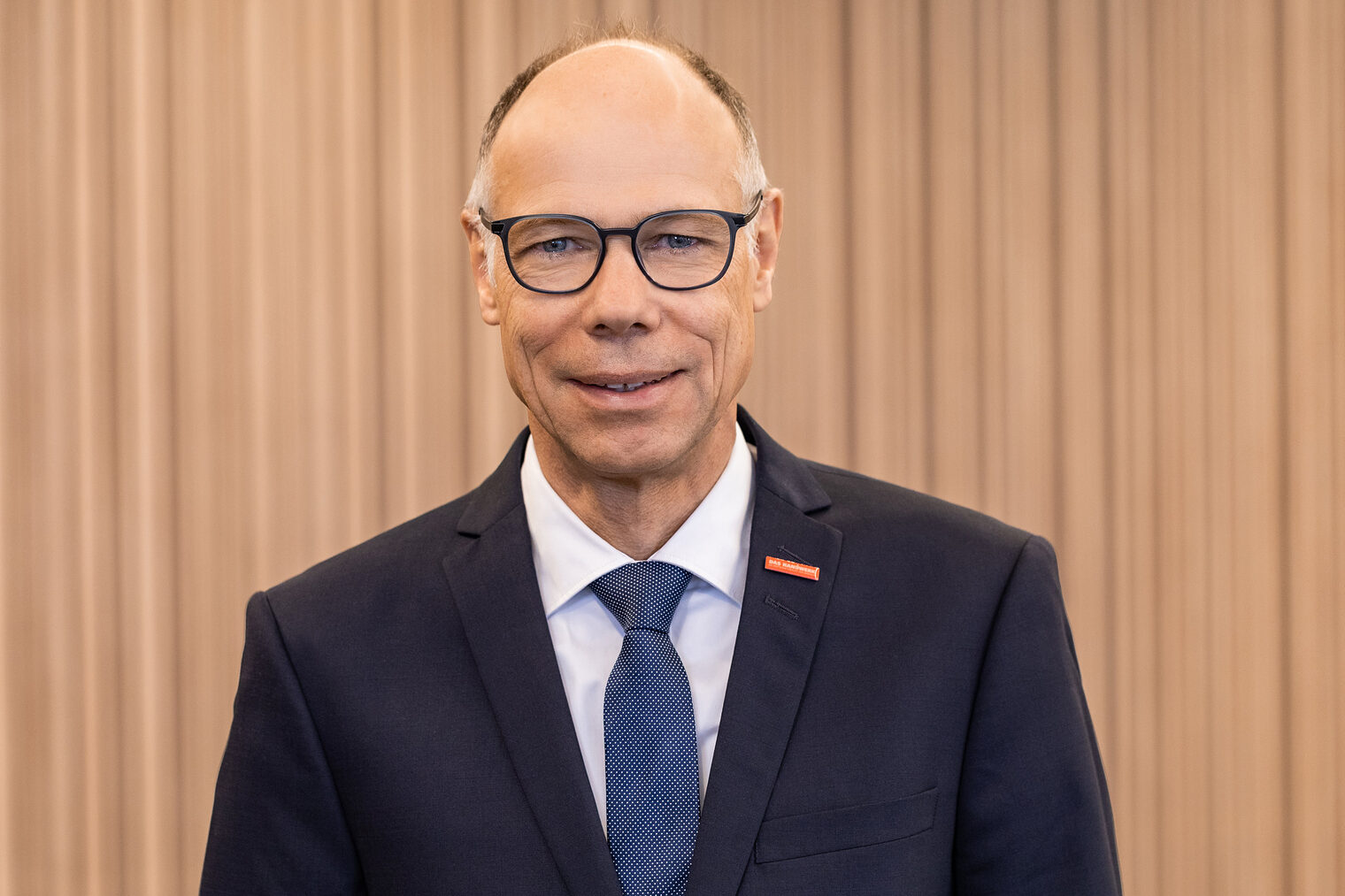 Mann mit dunkel gerahmter Brille und Glatze am oberen Kopf, trägt dunklen Anzug, weißes Hemd und blaue Krawatte, lächelt in die Kamera