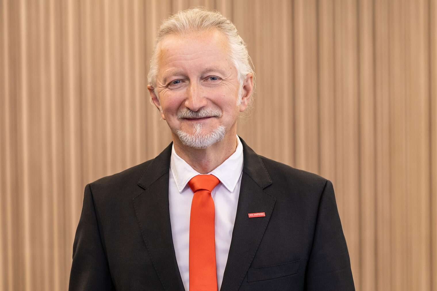 Mann Mit weißem, zurückgebundenem Haar und Oberlippen- und Kinnbart, dunkles Jackett, weißes Hemd und orangefarbene Krawatte, lächelt in die Kamera