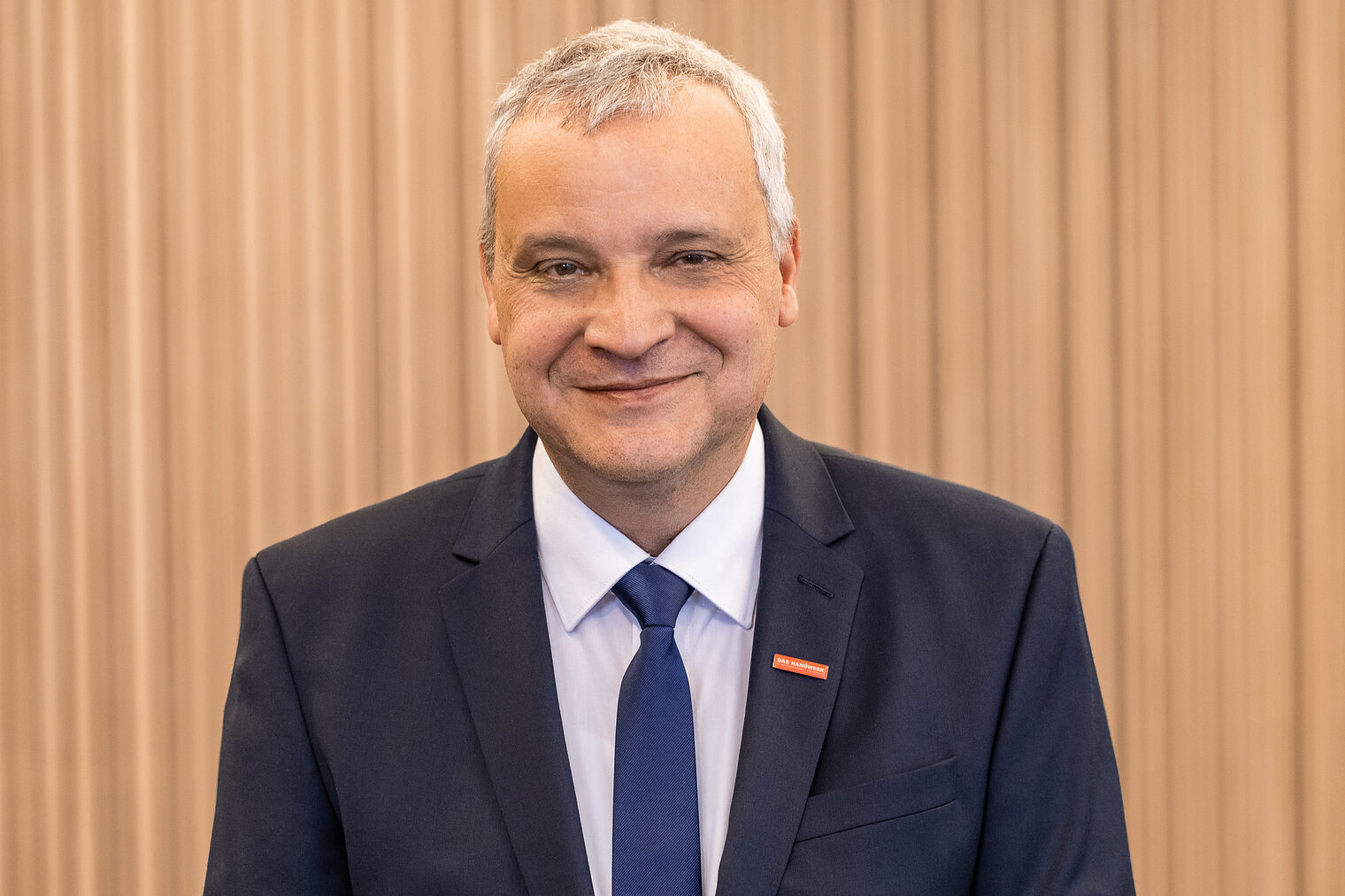 Mann mit glatt rasiertem Gesicht und weißem, kurzem, etwas schütterem Haar, dunkelblauem Jackett, weißem Hemd, blauer Krawatte, lächelt in die Kamera