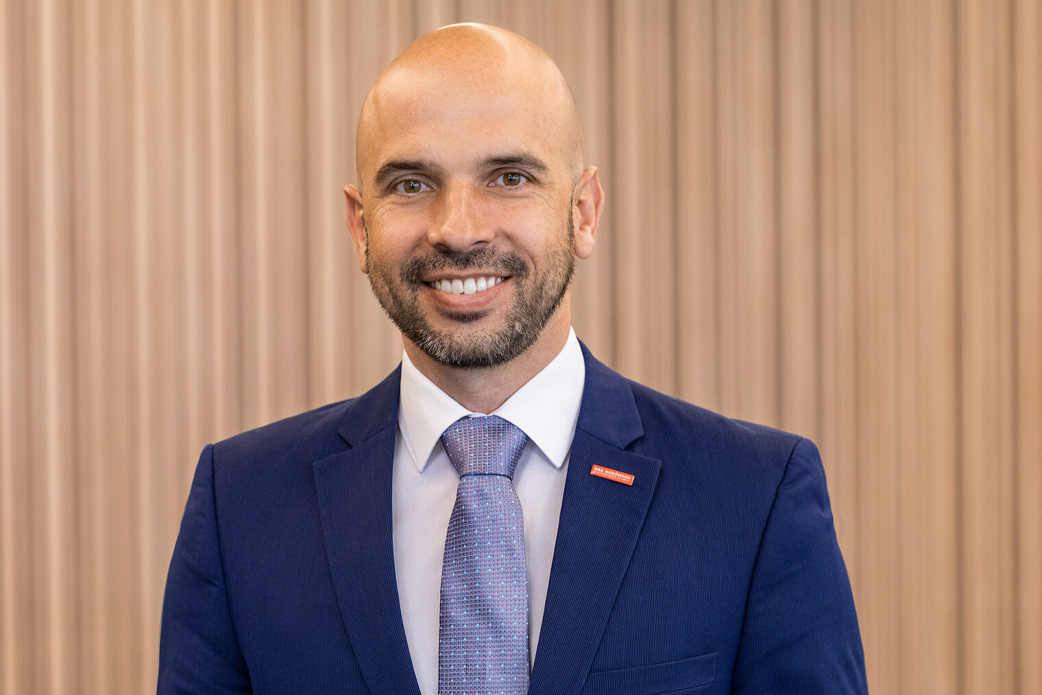 Mann mittleren Altes mit Vollglatze und dunklem, kurzem Vollbart, blaues Jackett, hellblaue Krawatte und weißes Hemd, lacht mit offenem Mund in die Kamera