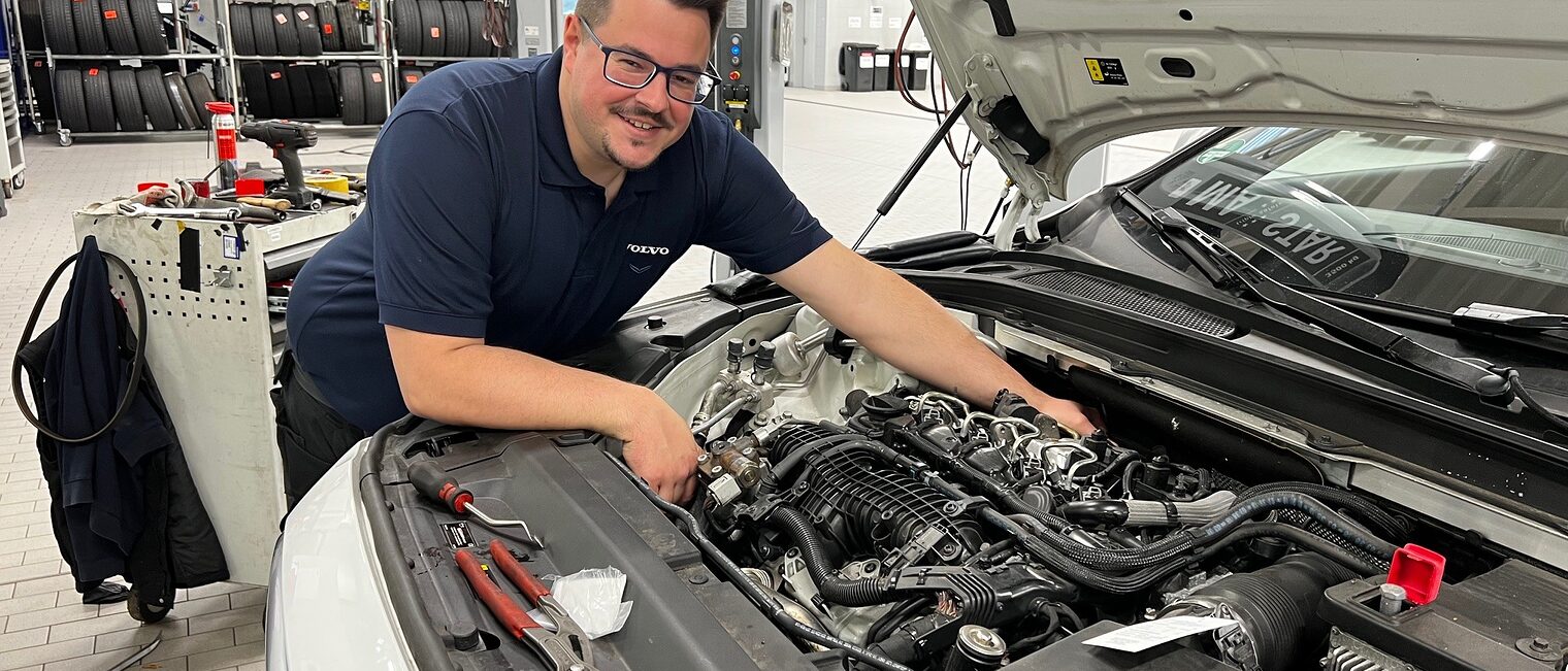 Handwerksmeister erzählen, warum die Weiterbildung so wertvoll ist Mann greift in Motorhaube von Auto und lächelt in die Kamera.