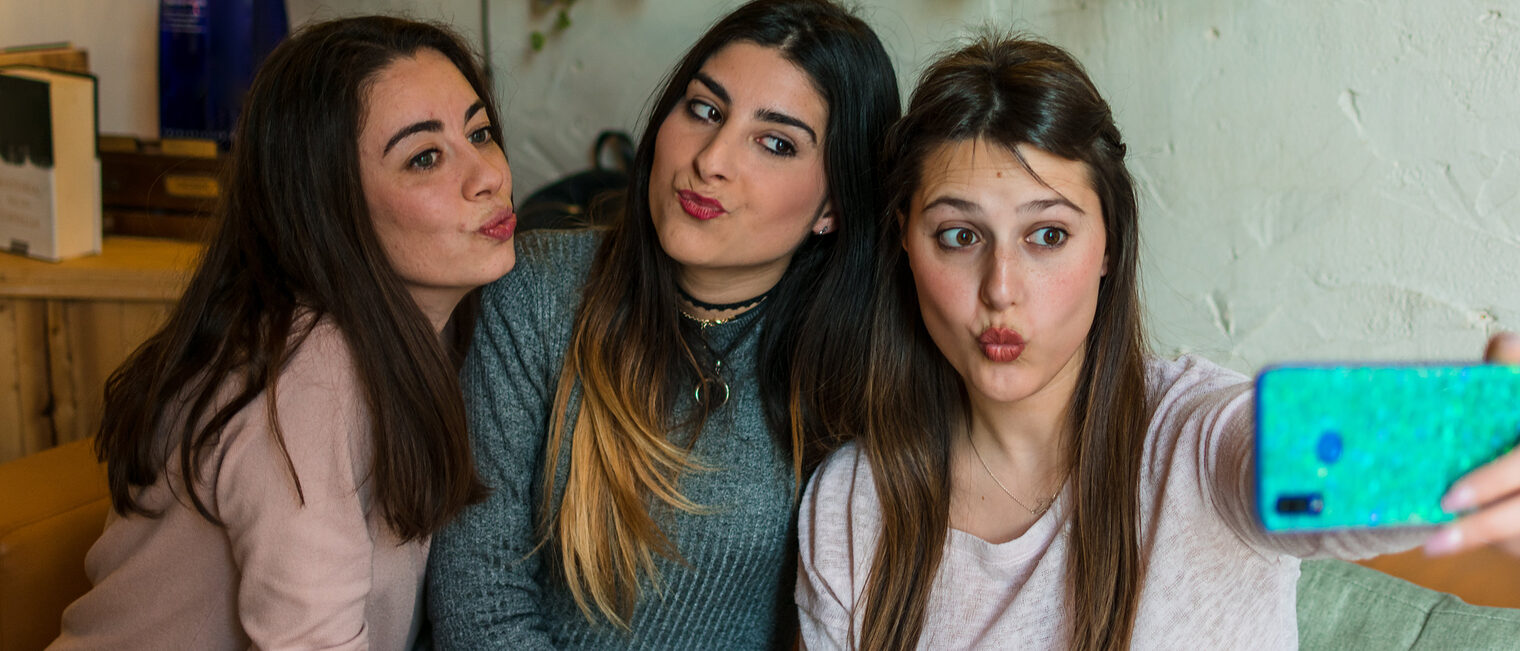 Drei jugendliche Frauen sitzen auf einem Sofa und posen in das Handy, das das rechte Mädchen für ein Selfie vor sich hält. Alle machen einen Kussmund.