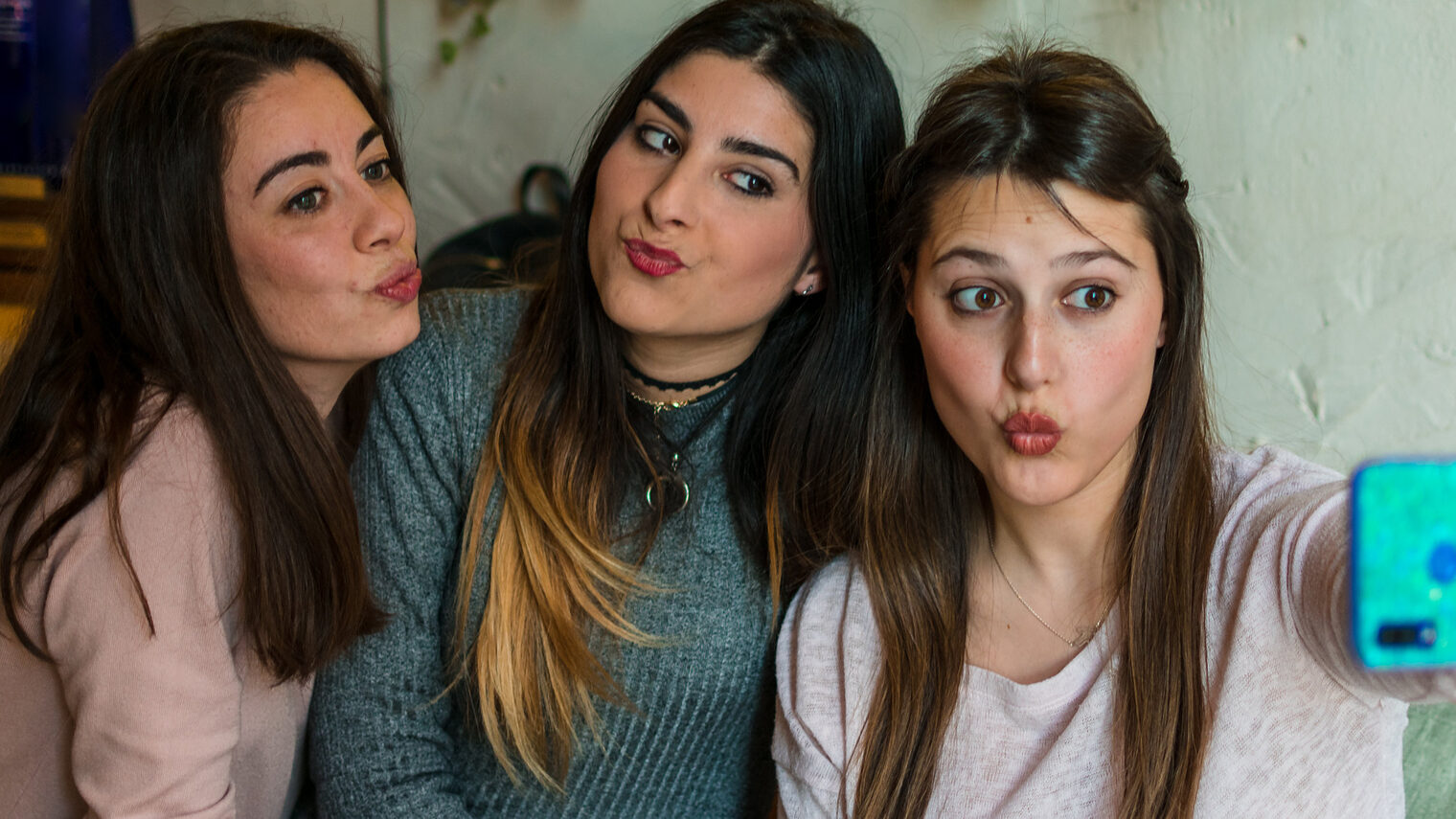 Drei jugendliche Frauen sitzen auf einem Sofa und posen in das Handy, das das rechte Mädchen für ein Selfie vor sich hält. Alle machen einen Kussmund.