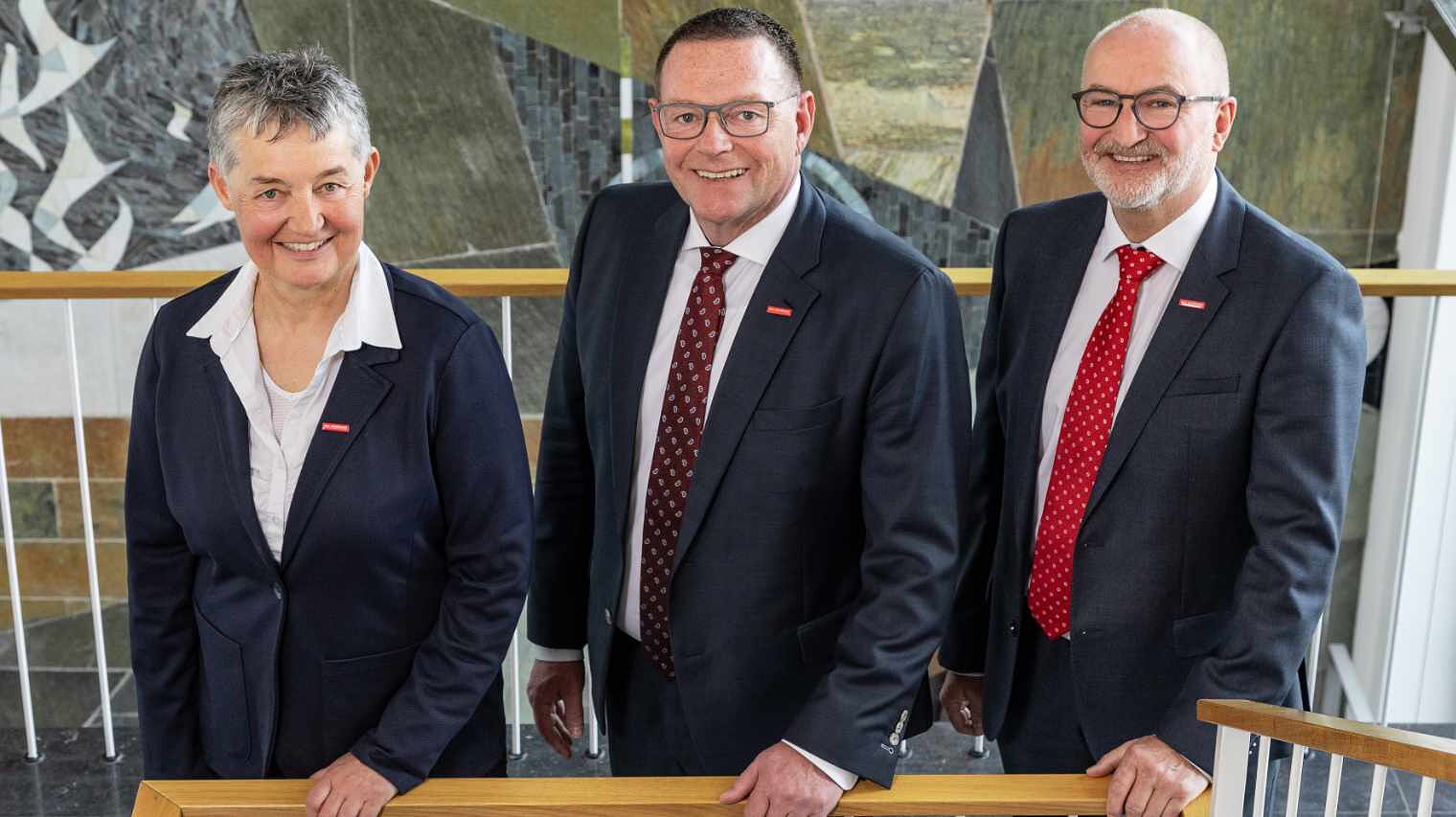 Drei Personen in offiziellen Outfits stehen im Trepphaus, ihre Hände auf dem Geländer und lächeln nach oben in die Kamera, links eine Frau, daneben zwei Männer