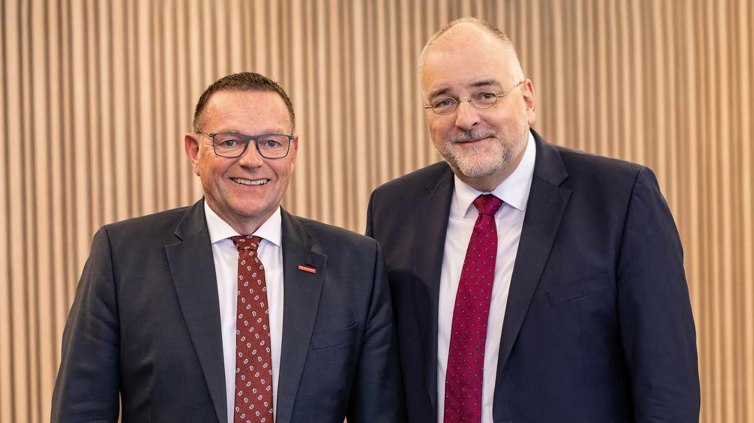 Zwei Männer mit Bille und dunklen Anzügen, weißen Hemden und roten Krawatten stehen nebeneinander und lachen in die Kamera, der rechte ist größer