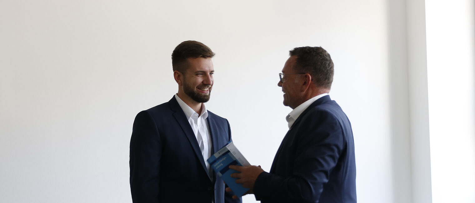 Kammerpräsident Klaus Hofmann bei Vereidigung von neuem Sachverständigen Mann in Anzug, der rechts steht, gratuliert jungem Mann links zum Amt des Sachverständigen.