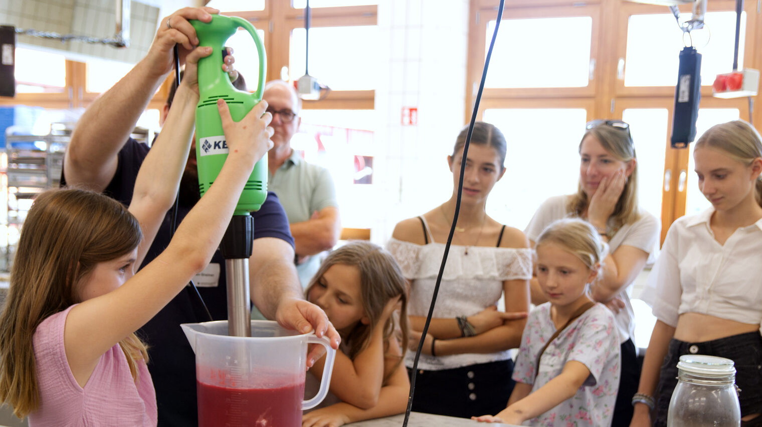Mädchen verschiedener Altersgruppen stehen in der Backstube um einen Tisch, auf dem mit einem riesigen Mixer rote Flüssigkeit gemixt wird.
