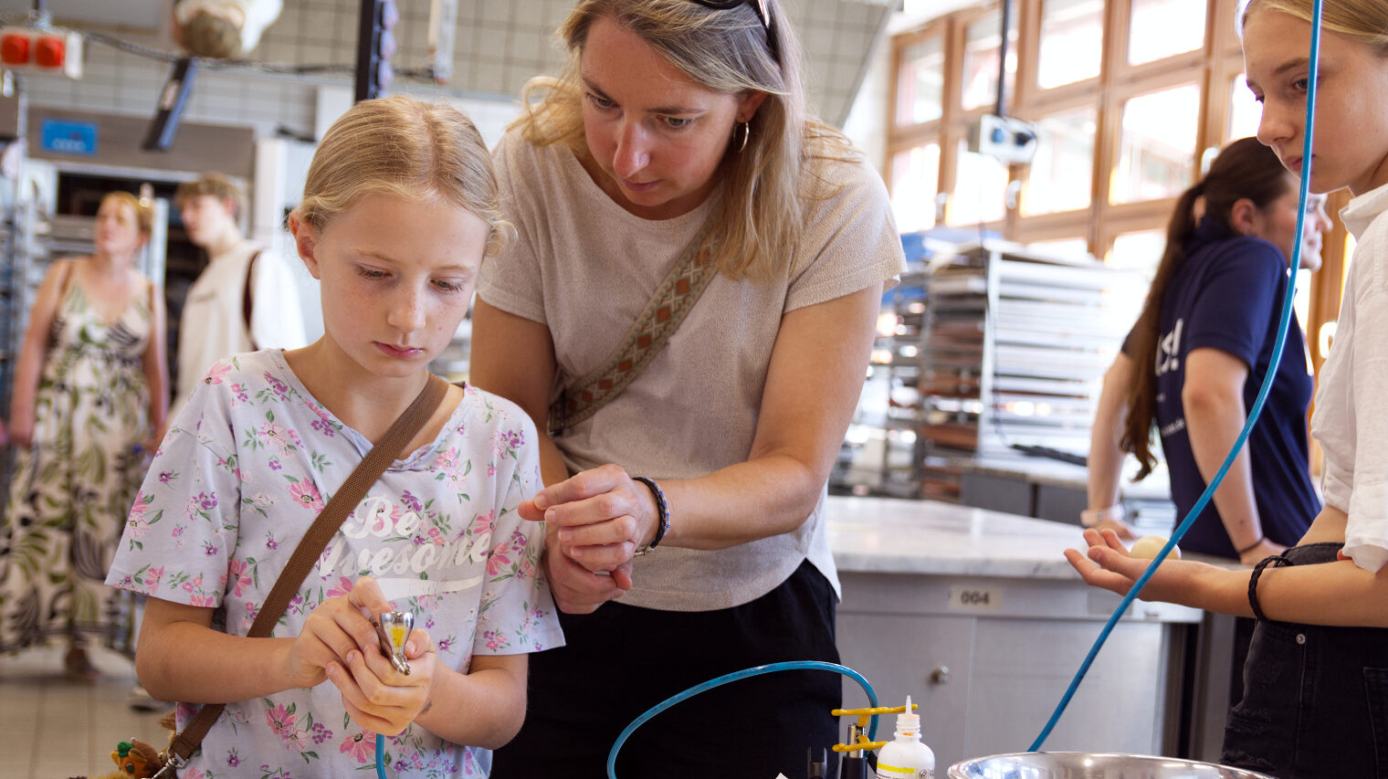 Mutter beugt sich über die Schulter eines kleinen blonden Mädchens und zeigt ihr in der Backstube wie den Umgang mit einer Sprühpistole