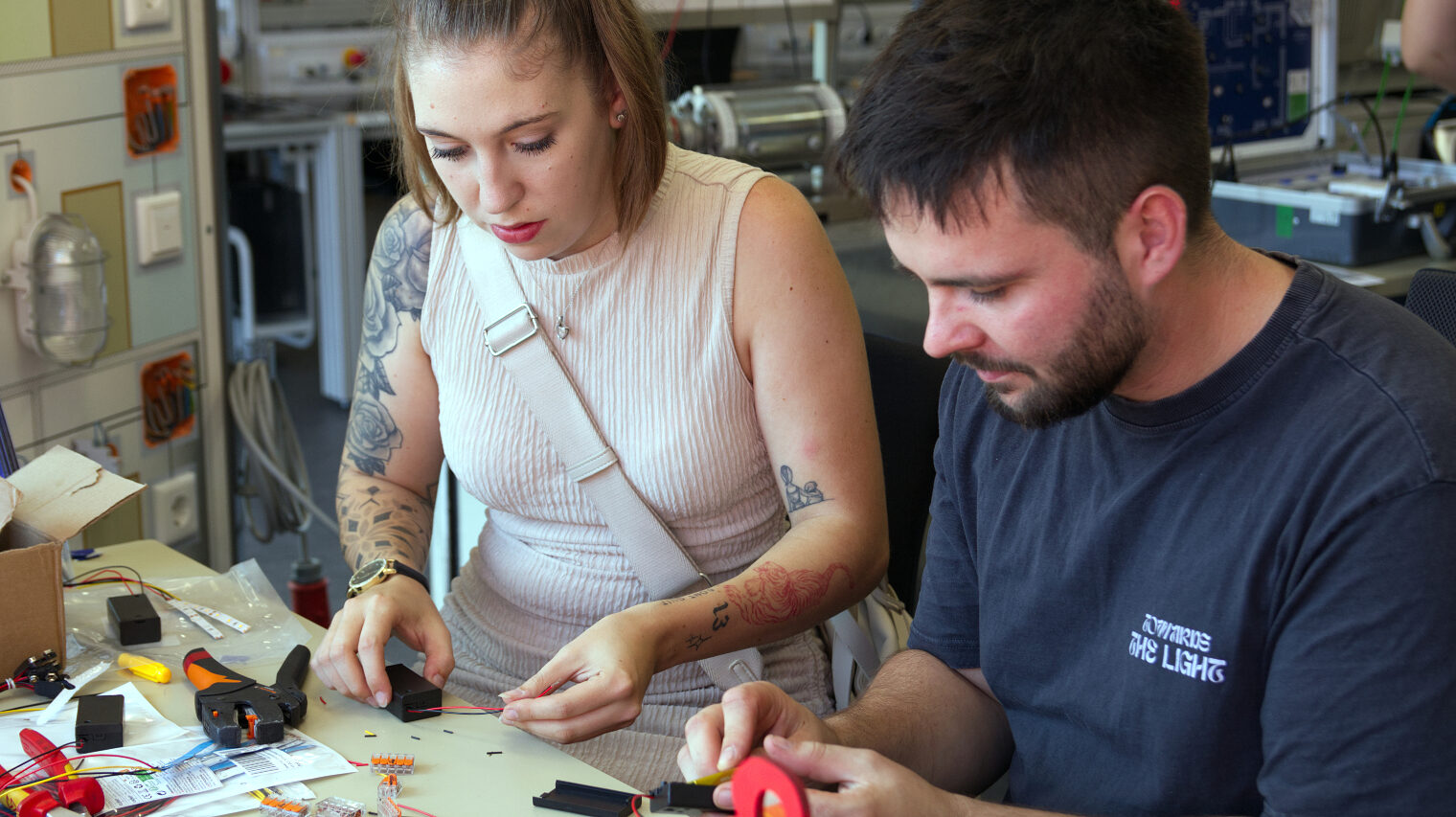Junge Frau mit tätovierten Armen und junger Mann sitzen gemeinsam an einen Werkstatt-Tisch und arbeiten mit Elektrokabeln