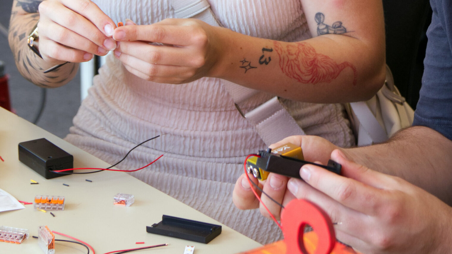 Detailansicht der Hände einer Frau mit tätovierten Armen und der Hände eines Mannes, die an einem Tisch Stecker verkabeln
