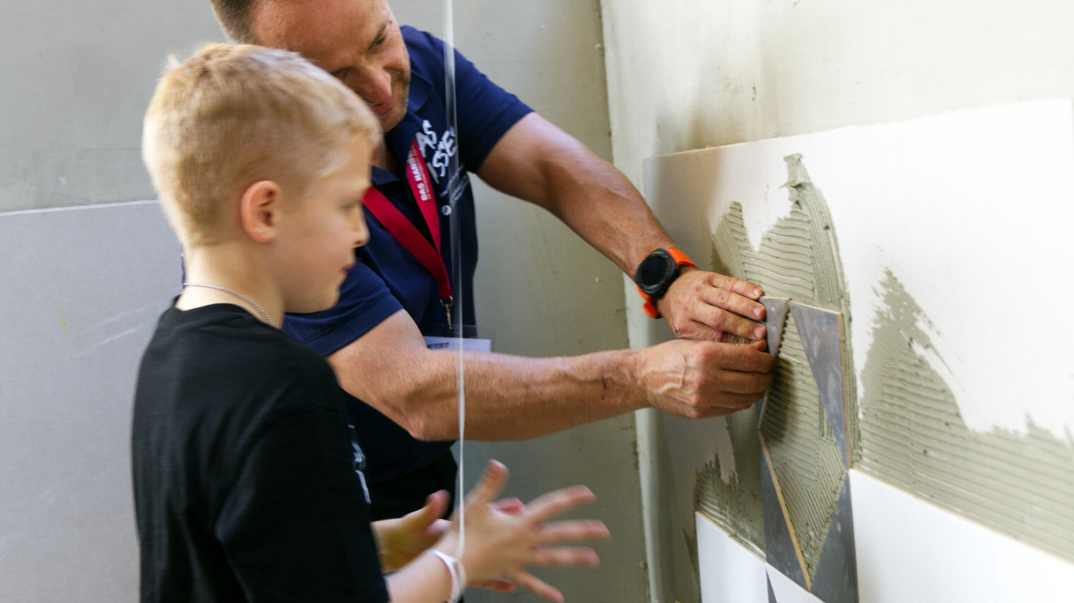 Handwerker zeigt einem Jungen wie man Fliesen an eine mit Mörtel bestrichene Wand klebt, der blonde Junge im Vordergrund schaut gespannt zu