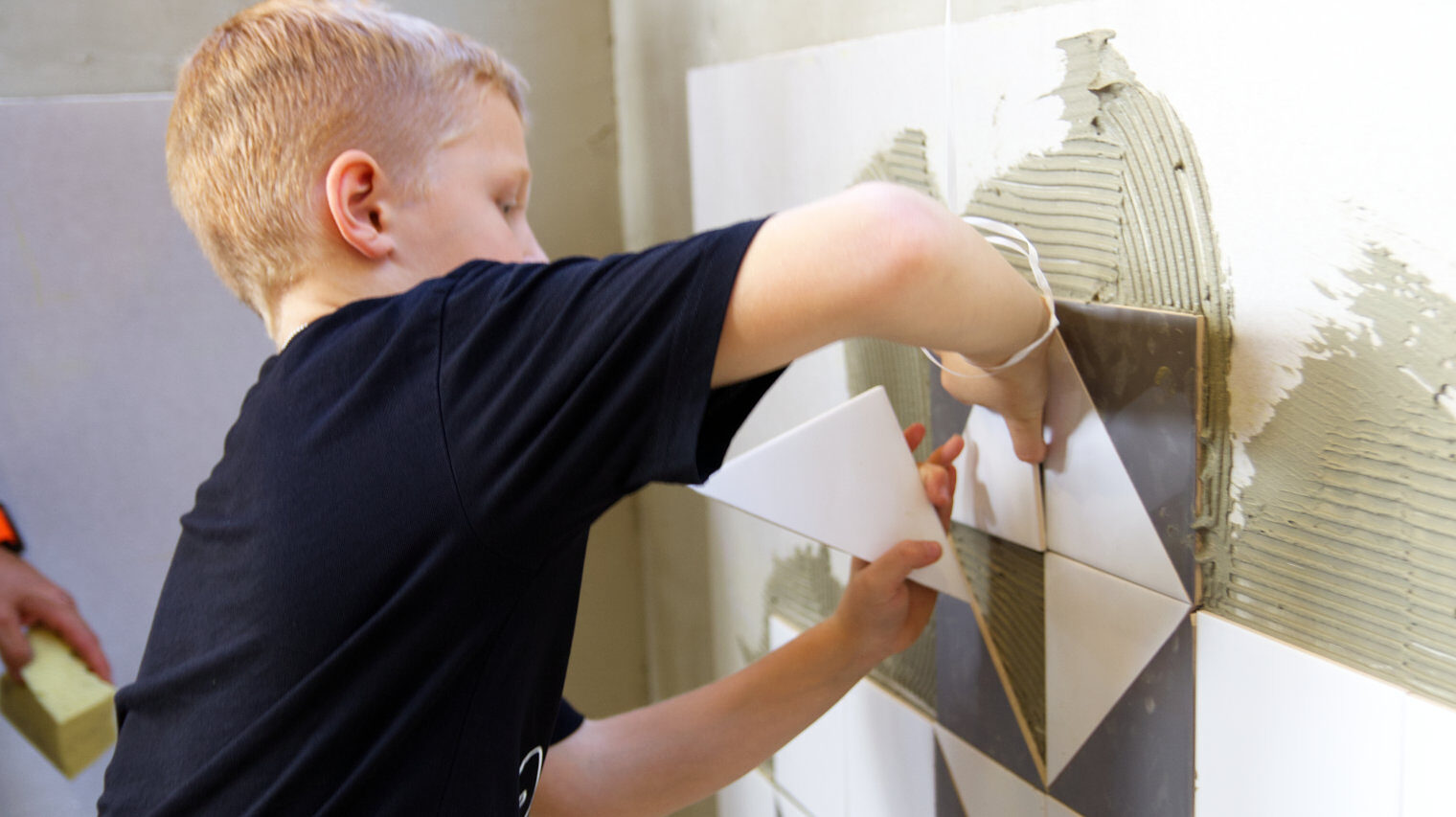 Blonder Junge klebt dreieckige Fliesen in Grau und Weiß zu einem Karomuster an eine mit Fliesenkleber bestrichene Wand