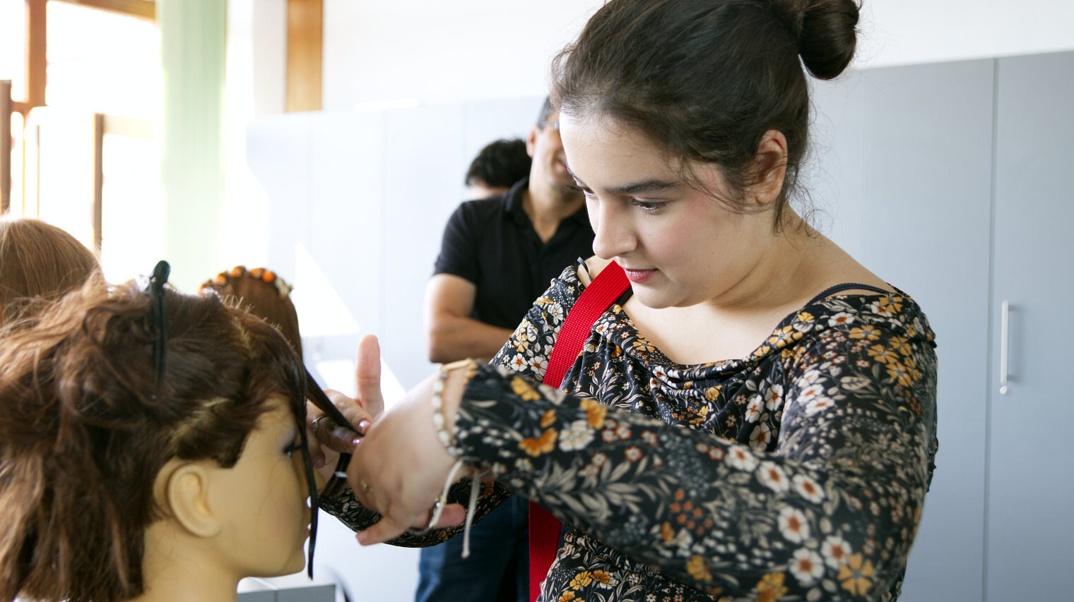 Junge Frau in geblümtem, langärmeligem Shirt mit zum Dutt locker hochgesteckten, braunen Haaren schneidet den Pony an einem Frisurenkopf