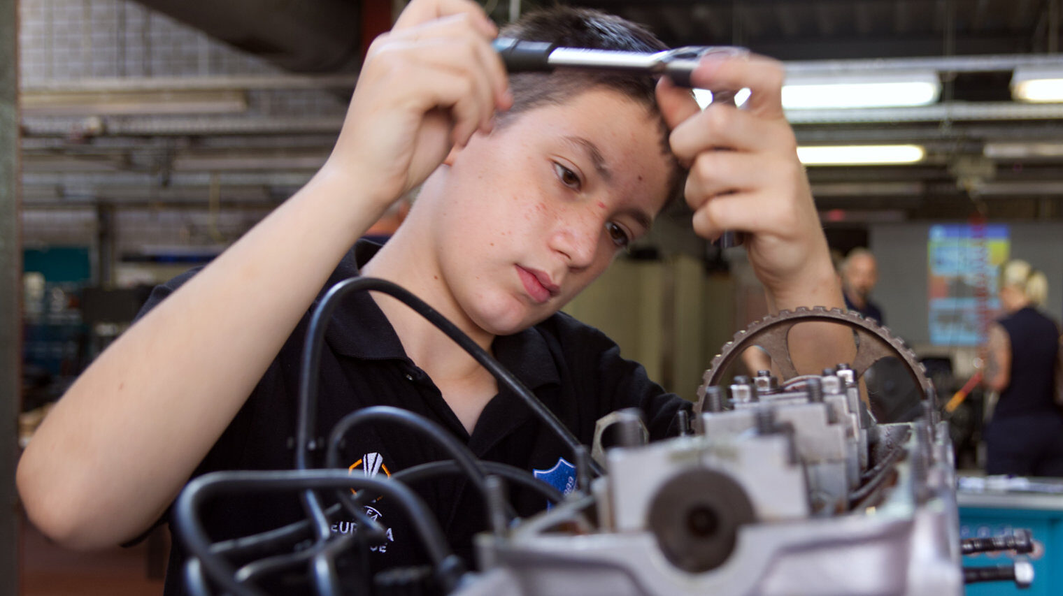 Ein Junge, vielleicht 14 Jahre, schraub an einem Motor im Bildvordergrund, dabei sind die Hände am oberen Bildrand und sein Kopf blickt dazwischen auf die Arbeit