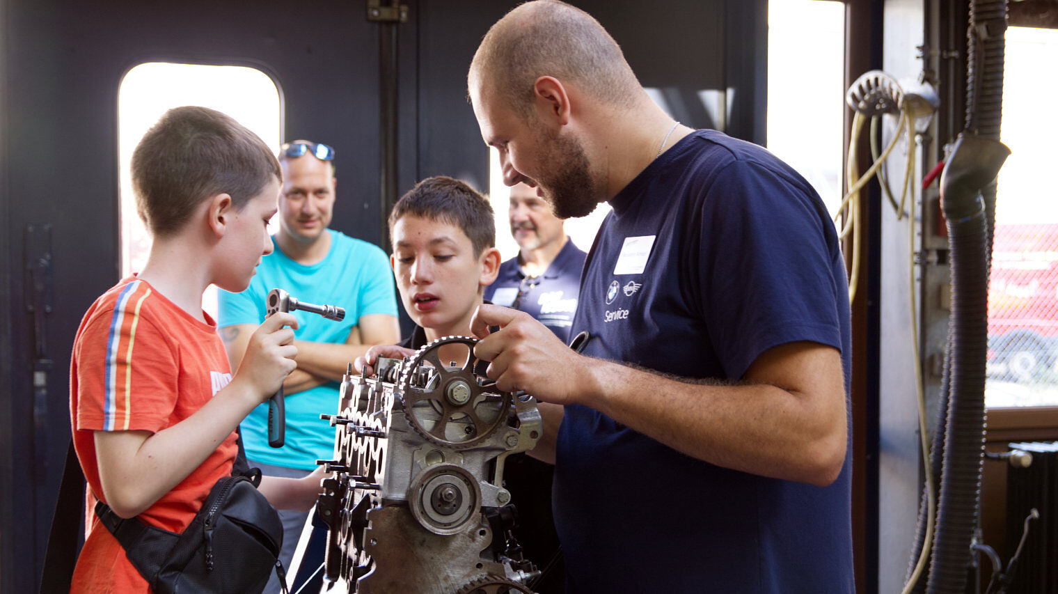 Um einen schon halb zerlegten Motor stehen Personen, links ein Junge im roten Shirt mit einem Schrauber in der Hand, dahin der staunende Kopf eines anderen Jungen, rechts ein Mann mit Bart, im Hintergrund unscharf zwei weitere Männer