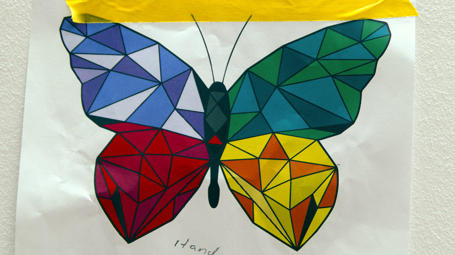 Papier mit der Zeichnung eines Schmetterlings, die mit einem gelben Klebestreifen an die Wand geklebt ist, die Flügel des Schmetterlings sind zu Dreiecken unterteilt in verschiedenen Farben ausgemalt