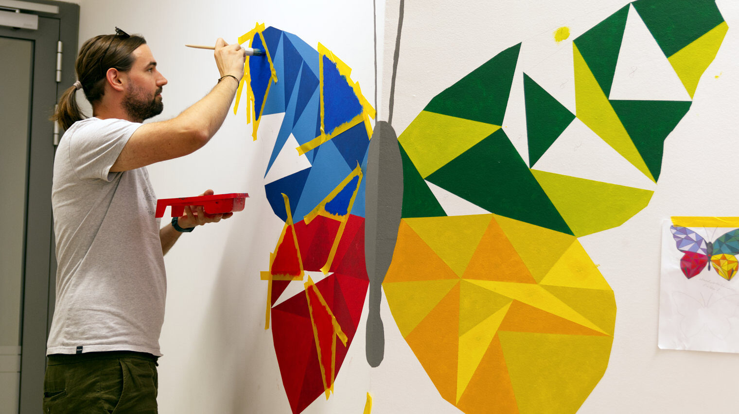 Mann mit Zopf im weißen Shirt vor einer Wand, auf der ein großer Schmetterling in verschiedenen Farben entsteht, mit der rechten Hand bemalt er eine Fläche, in der linken hält er die Farbe