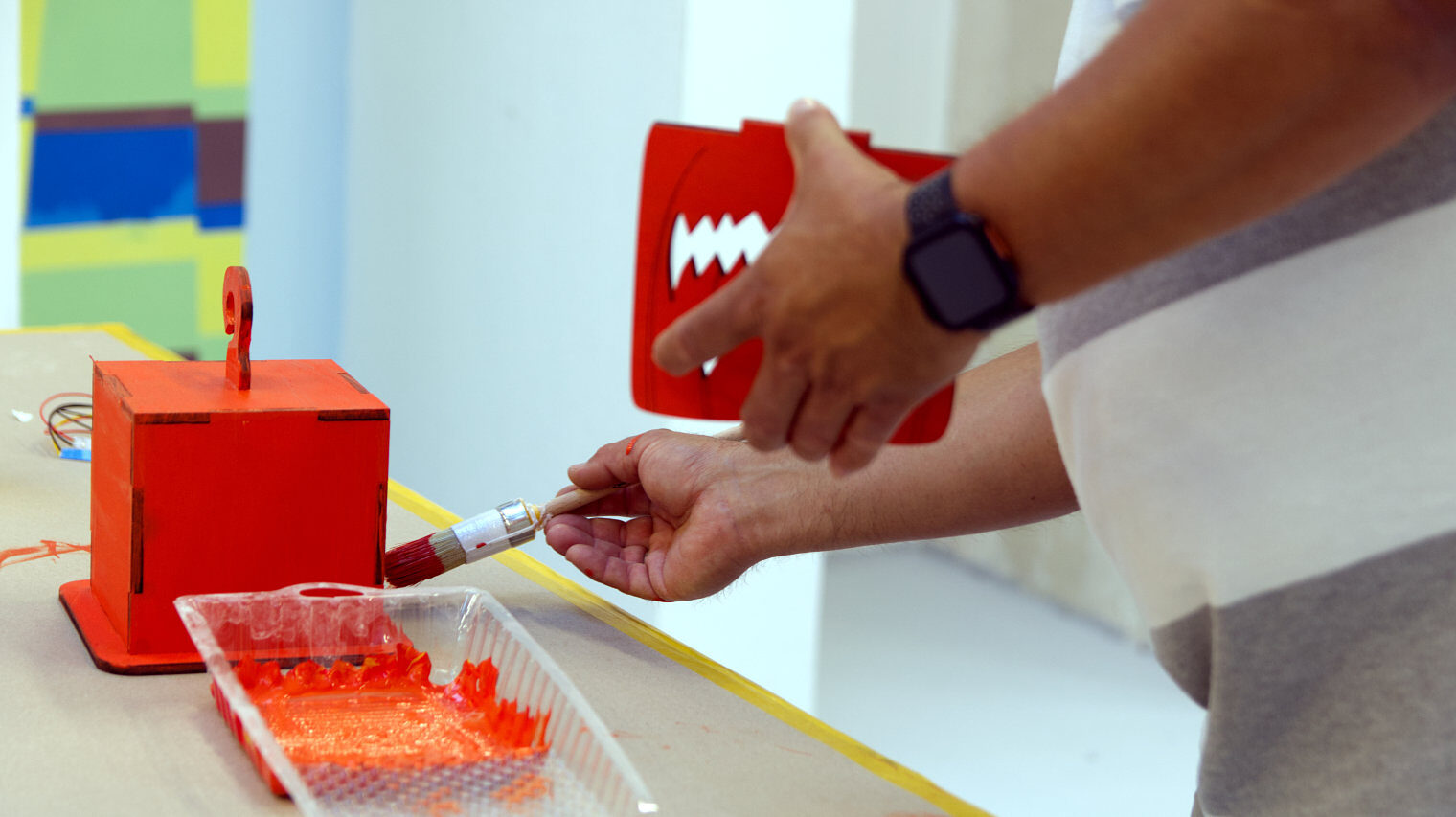 Nahansicht einer Streicharbeit, vorne die Palette mit orangener Farbe, dahinter ein Kubus, eine Hand führt einen Pinsel darüber, in der anderen Hand hält der Mann die Vorderseite des Kubus mit Kürbisgeistgesicht