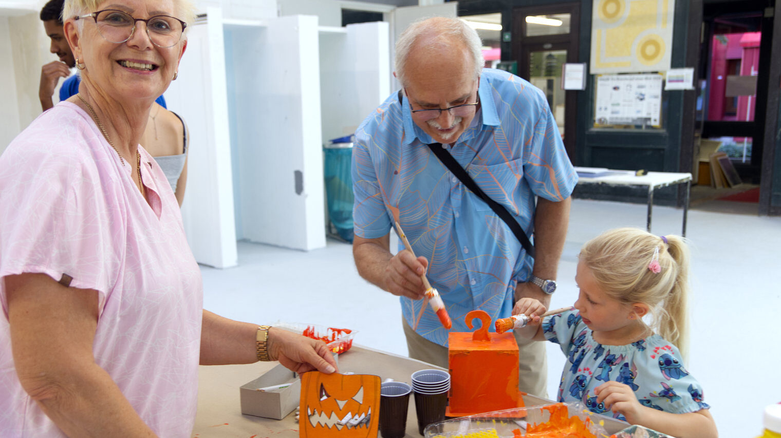 Links eine ältere Frau mit kurzen weißen Haaren, Brille und rosa Shirt, die das bemalte Kürbisgeist-Gesicht in die Kamera zeigt, rechts bemalen Opa und kleine blonde Enkelin den Kürbiskörper in Orange