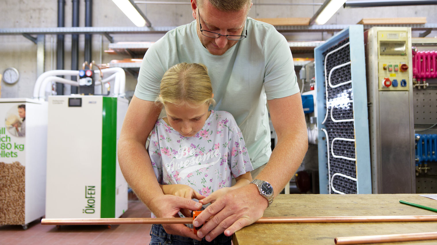 Blick in die SHK-Werkstatt, mittig im Bild ein Vater mit Brille, der sich von hinten über ein kleines blondes Mädchen beugt, um mit ihm gemeinsam ein Metallrohr zu durchschneiden