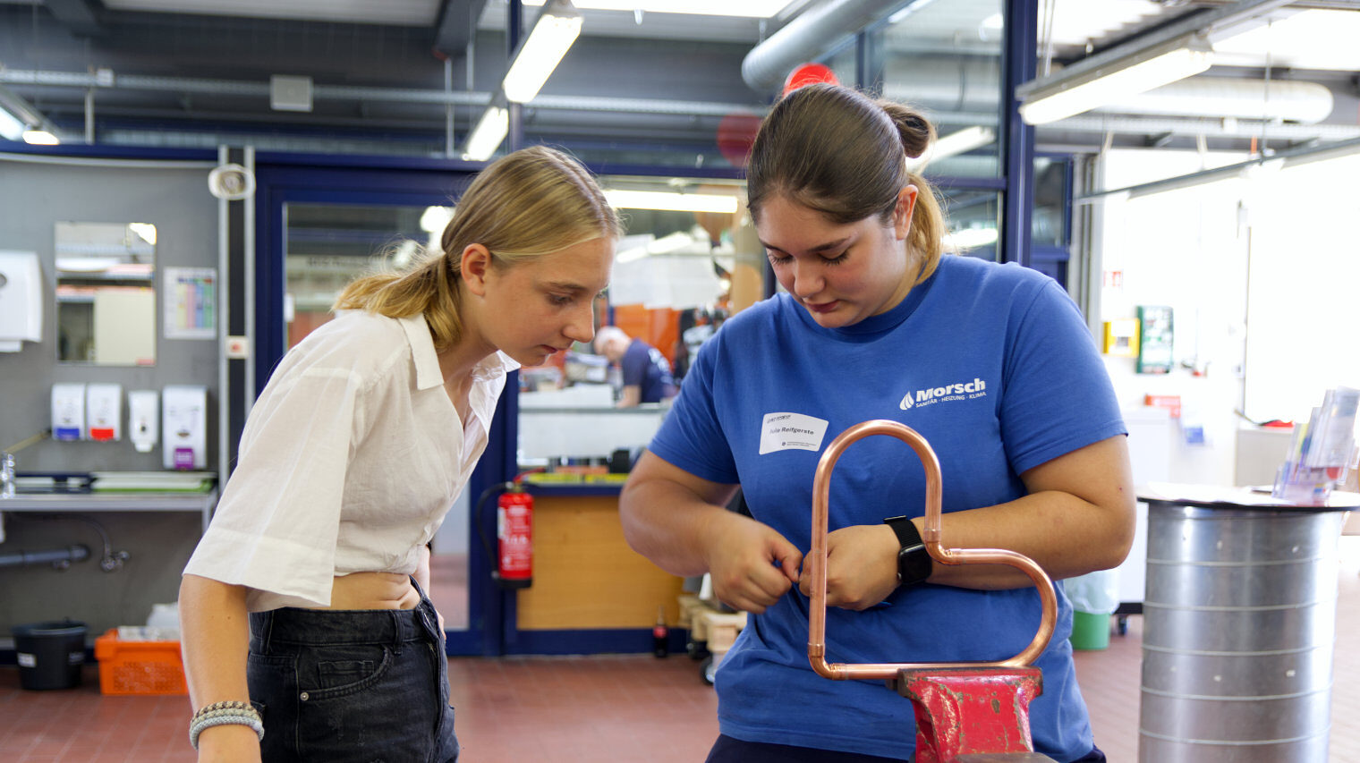 Zwei junge Frauen in einer Werkstatt, die rechte arbeitet in ihren Handen etwas aus, die linke schaut nach vorne gebückt genau zu, im Vordergrund ist ein Herz aus Metall eingespannt