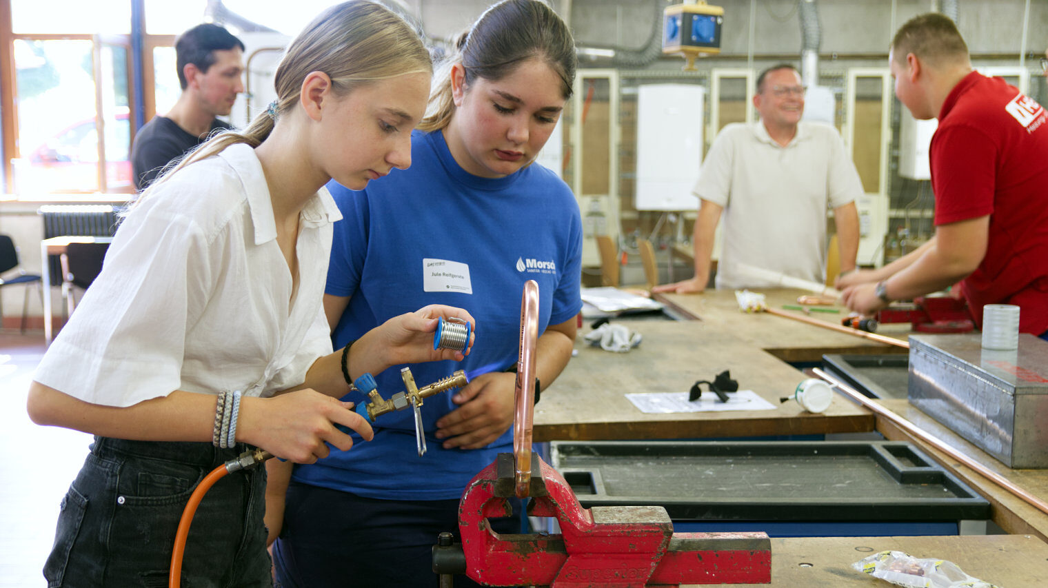 Blick in die SHK-Werkstatt mit zwei Mädchen im Vordergrund, die Blonde verlötet ein in den Schraubstock eingespanntes Metallstück, die andere schaut zu, im Hintergrund unscharf Männer im Gespräch