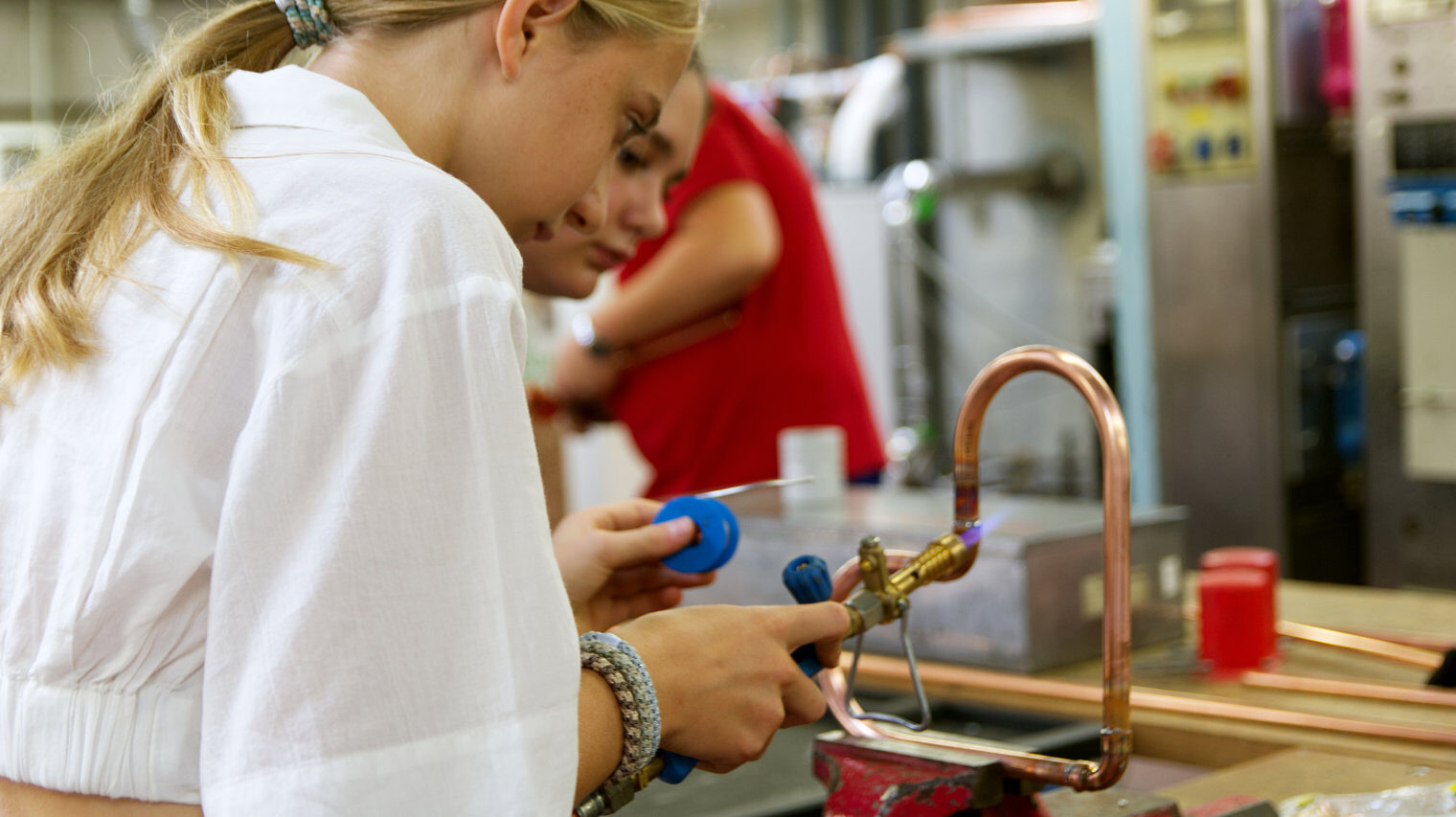 seitliche Ansicht eines Mädchens mit blondem Haarzopf und weißer Bluse, die in einer Werkstatt ein Herz aus Metallrohr verlötet, leicht verdeckt im Hintergrund schaut ein zweites Mädchen zu