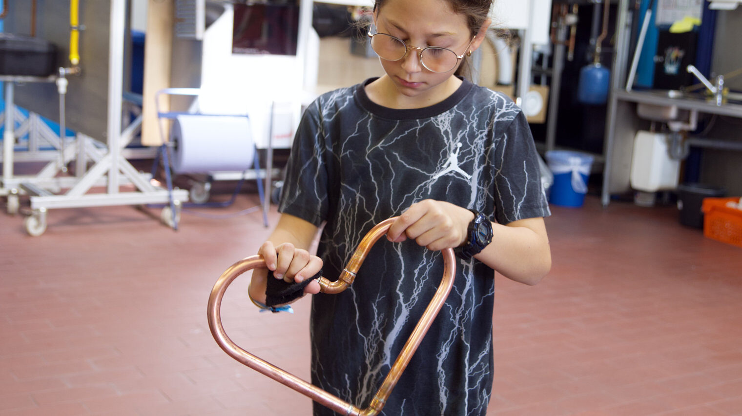 Kleines Mädchen mit großer Brille und braunem Haarzopf in einem Ausbildungsraum hält ein Herz aus Metallrohr in der Hand und blickt darauf, während sie es poliert