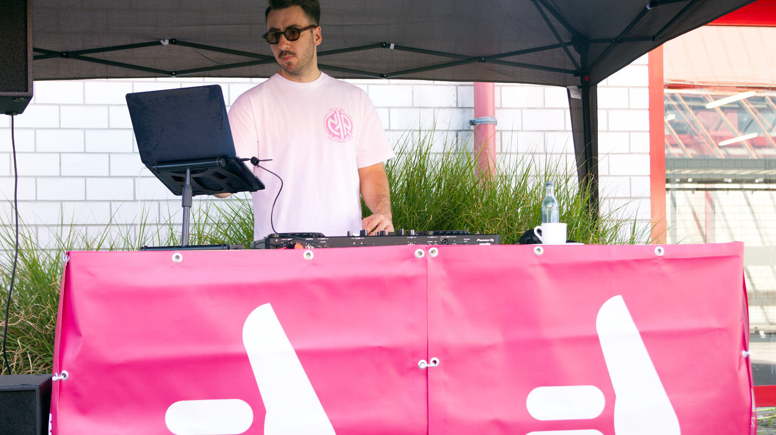 DJ mit dunkler Sonnenbrille und dunklen kurzen Haaren steht im weißen T-Shirt an seinem Mischpult unter einer Plane im Freien, davor sind stilisierte Daumen auf einer Plane zu sehen