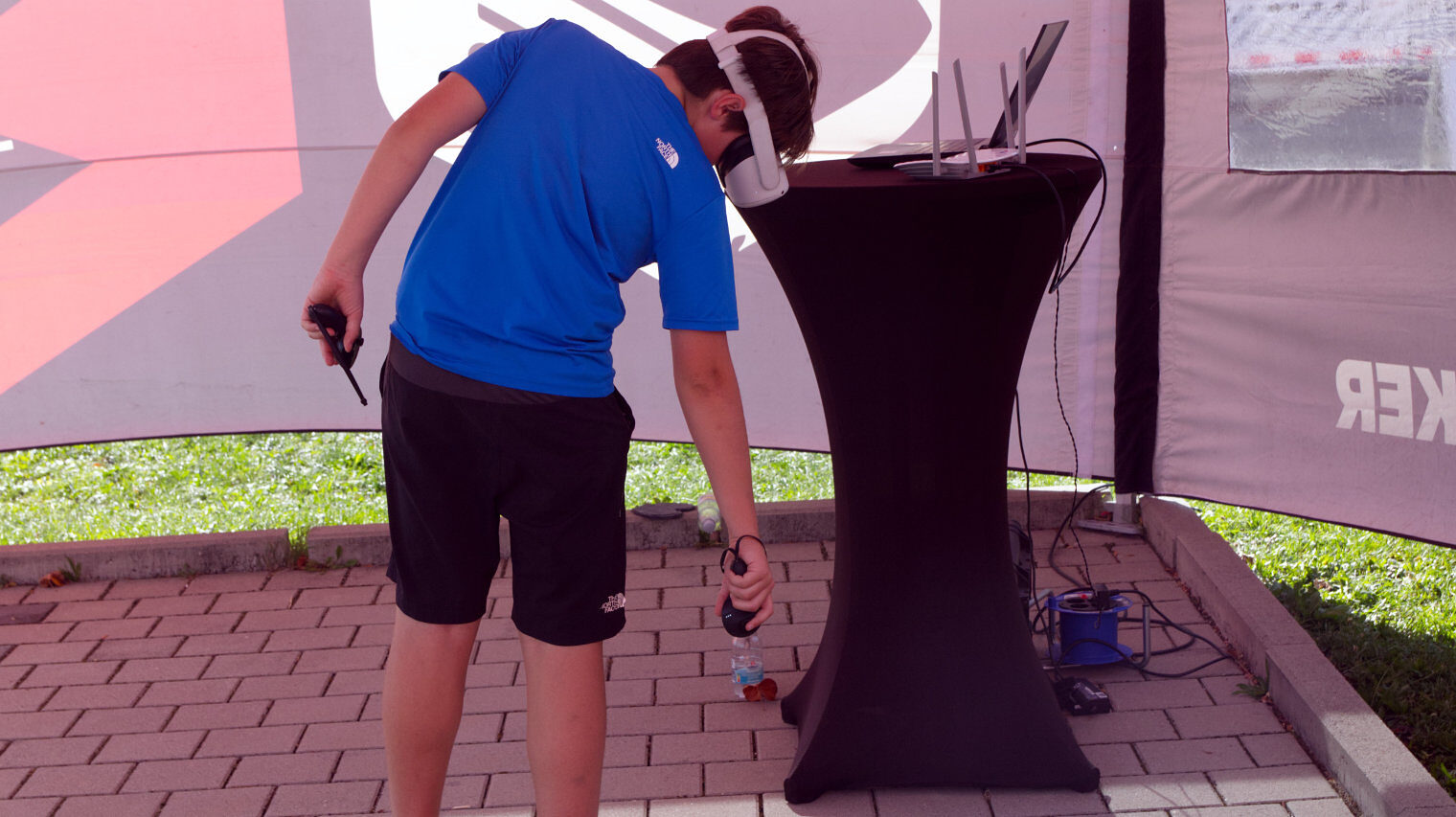 Junge in kurzen Hosen und blauem T-Shirt mit einer VR-Brille vor dem Gesicht agiert in einem Zelt draußen vor einem Stehtisch mit dem Controller und zeigt dabei zu Boden
