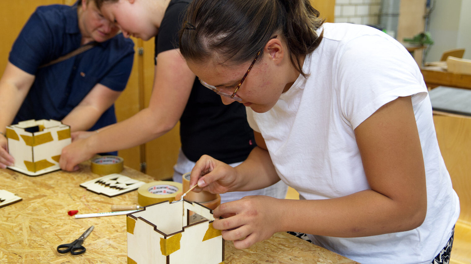 Mädchen mit dunklen, Haaren, Brille und weißem T-Shirt beugt sich über einen Holzwürfel und verleimt die Kanten, im Hintergrund arbeiten 2 weitere Frauen ebenso