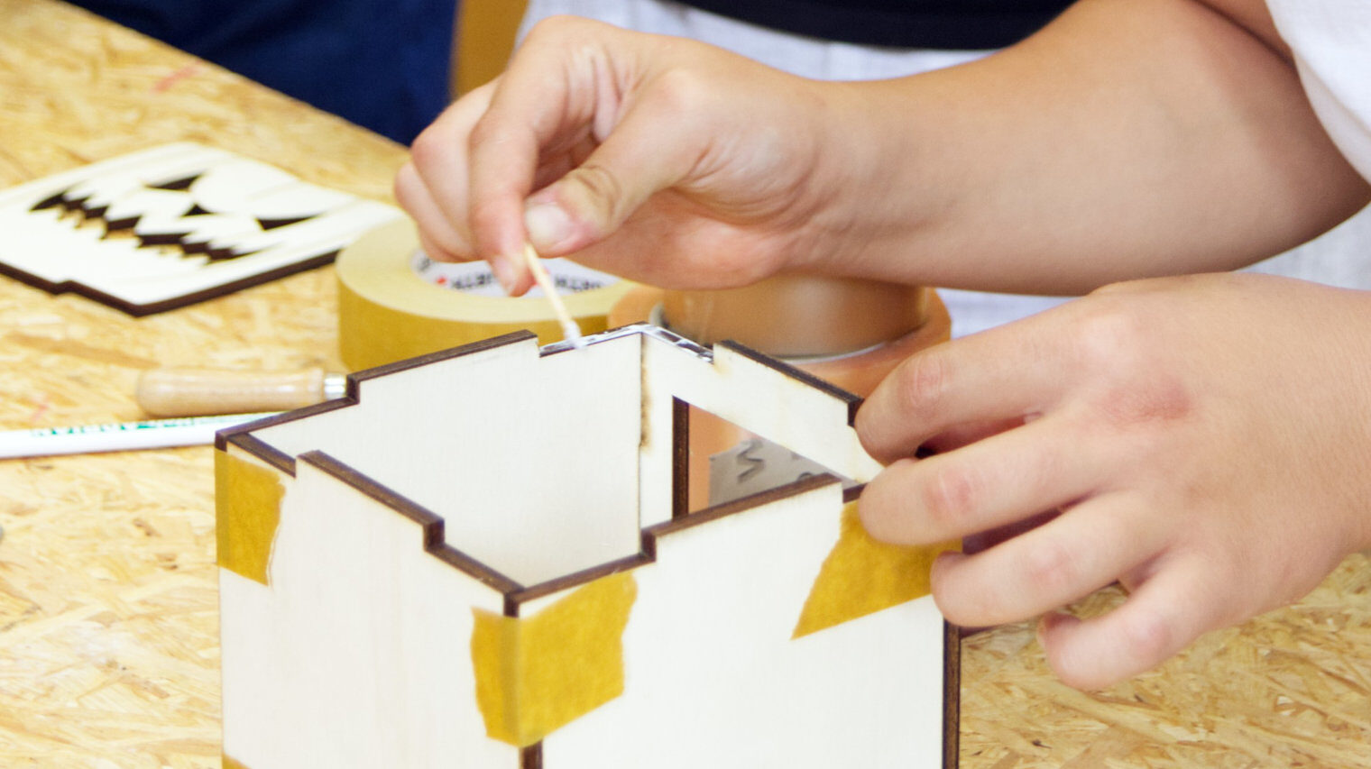 Detailaufnahme von jungen Händen, die an einem Holzwürfel mit einem kleinen Stäbchen Kleber auf die Kanten auftragen, auf dem Tisch liegen Kreppband und ein Kürbisgesicht aus Holz