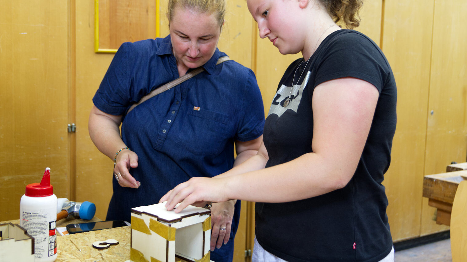 Zwei Frauen an einem Tisch vor einer Holzschrankwand in einer Werkstatt, arbeiten gemeinsam an einem Holzwürfel, der verleimt wird, auf dem Tisch weitere Arbeitsmaterialien