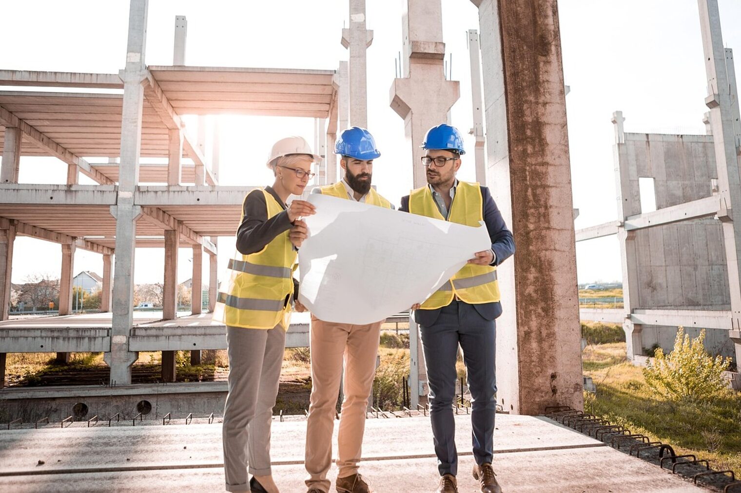 Im Hintergrund befindet sich eine Baustelle eines Gebäudes, dass sich im Rohbau befindet. Im Vordergrund stehen drei Männer mit Warnerste und Helm, die alle auf einen Bauplan gucken. 
